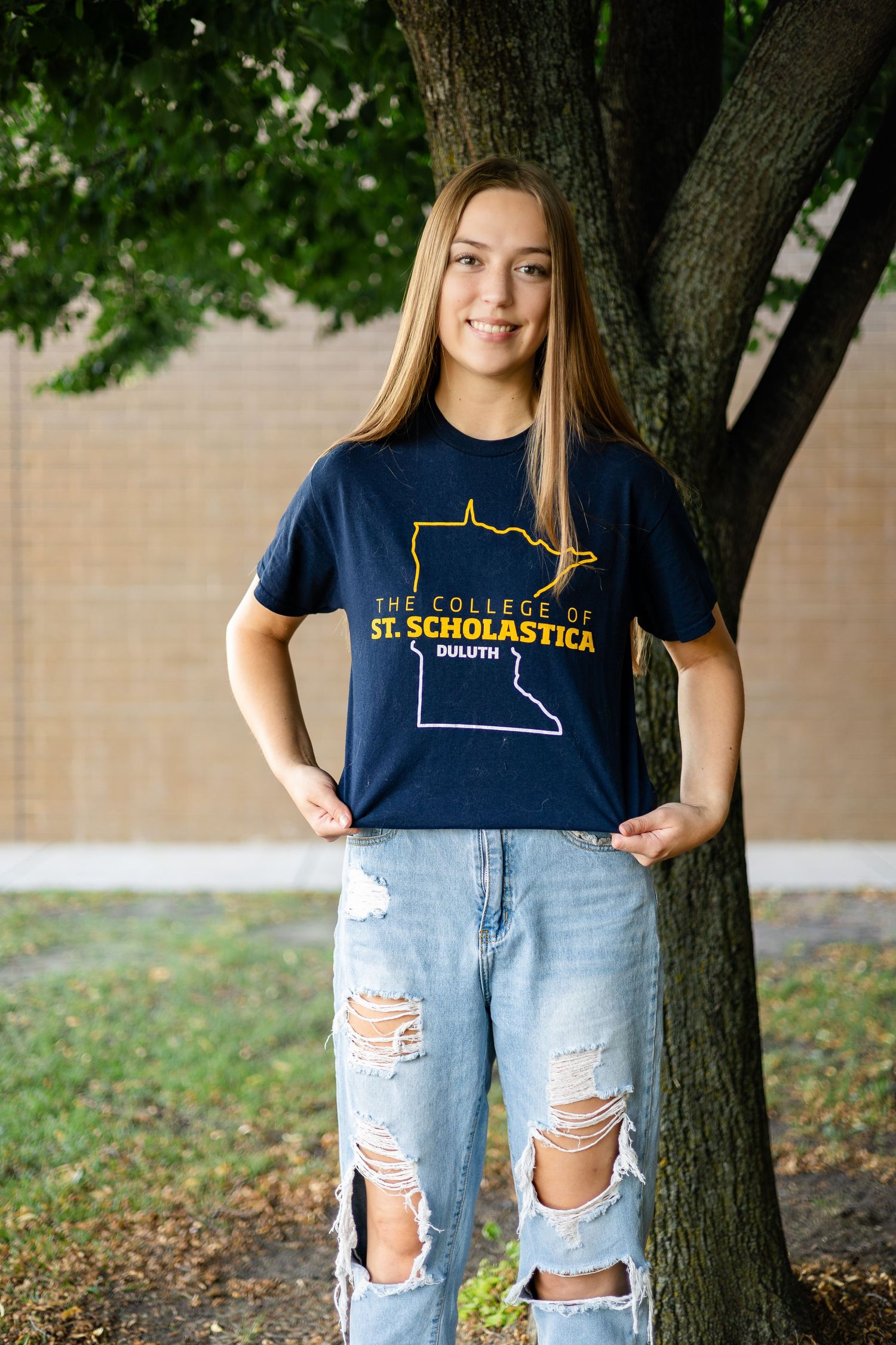 Student wearing St. Francis High School t-shirt and ripped jeans poses against tree.