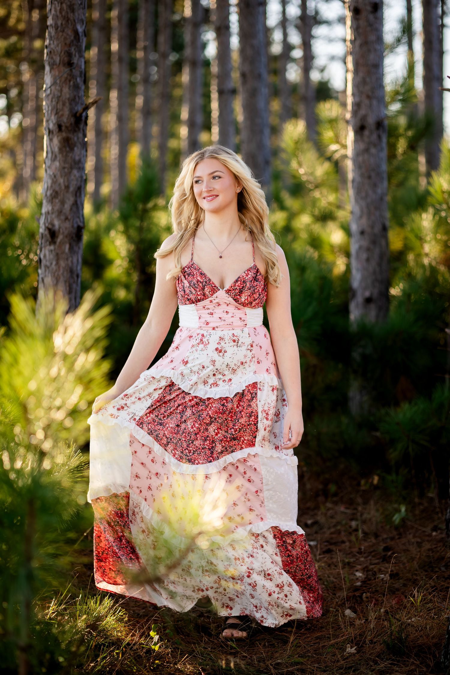Woman wearing flowy red and white dress poses in sunlit forest setting.