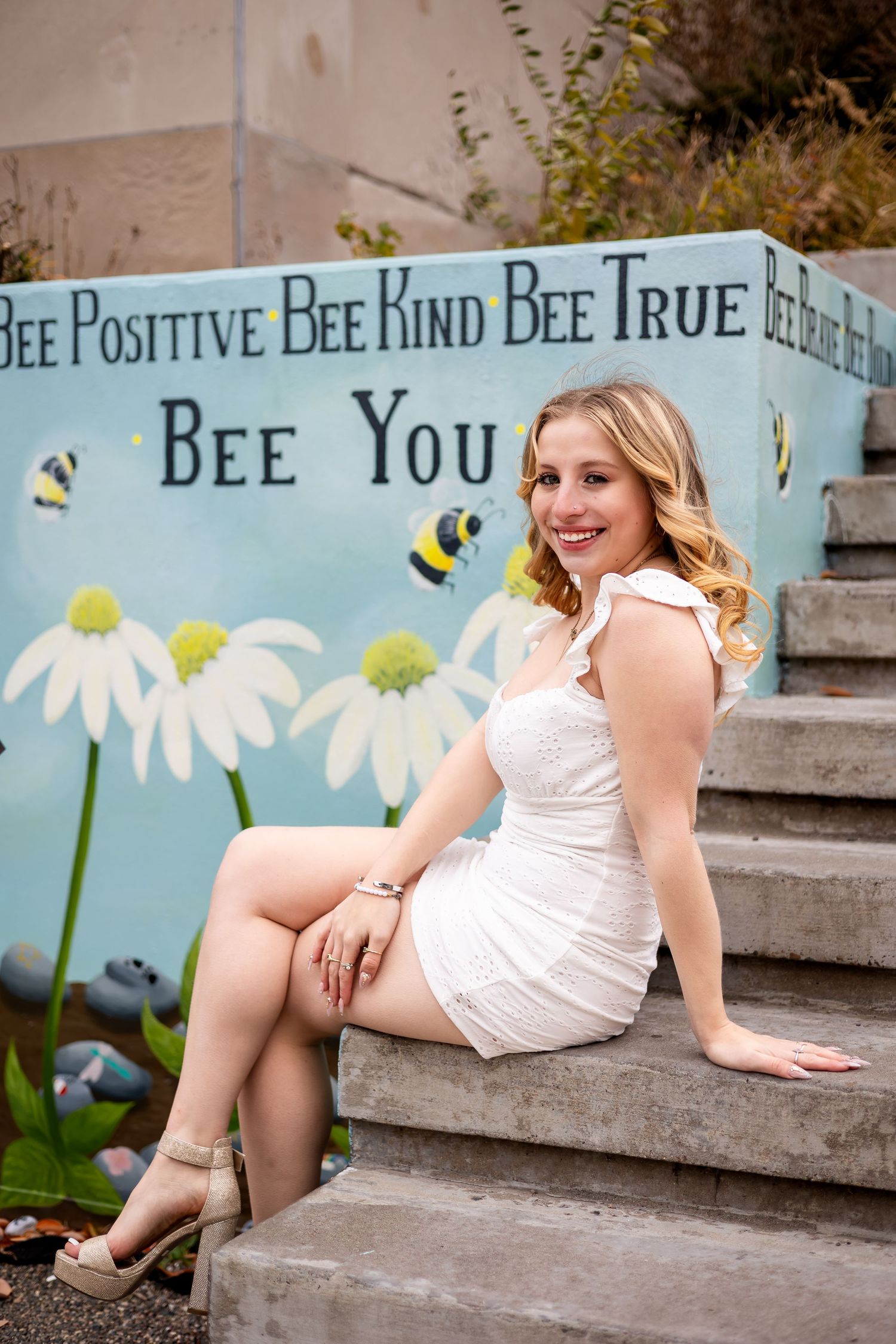 A person in a white dress poses on concrete steps against a painted wall with bees and daisies.