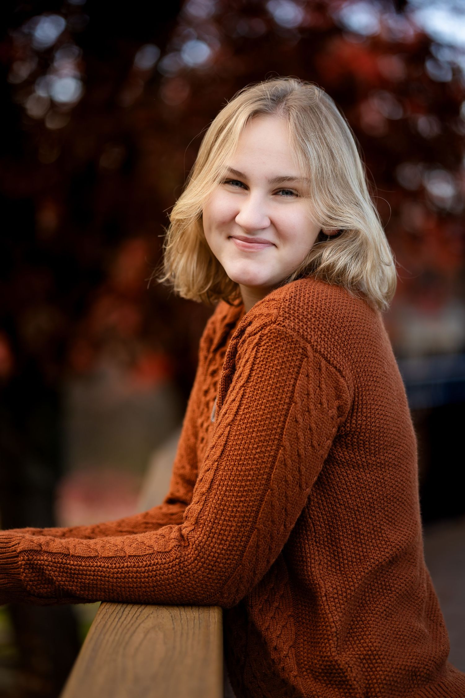 A person wearing a cozy rust-colored knit sweater poses outdoors against an autumn background.