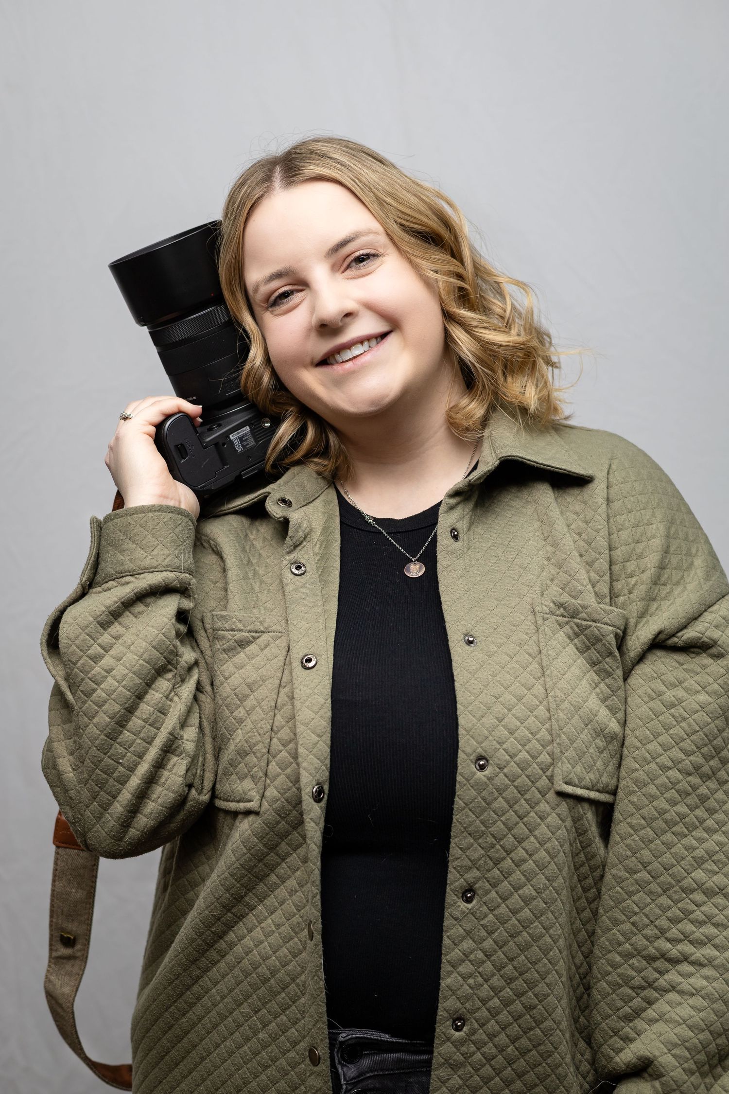 A photographer in an olive green quilted jacket holds a professional camera against a gray studio background.
