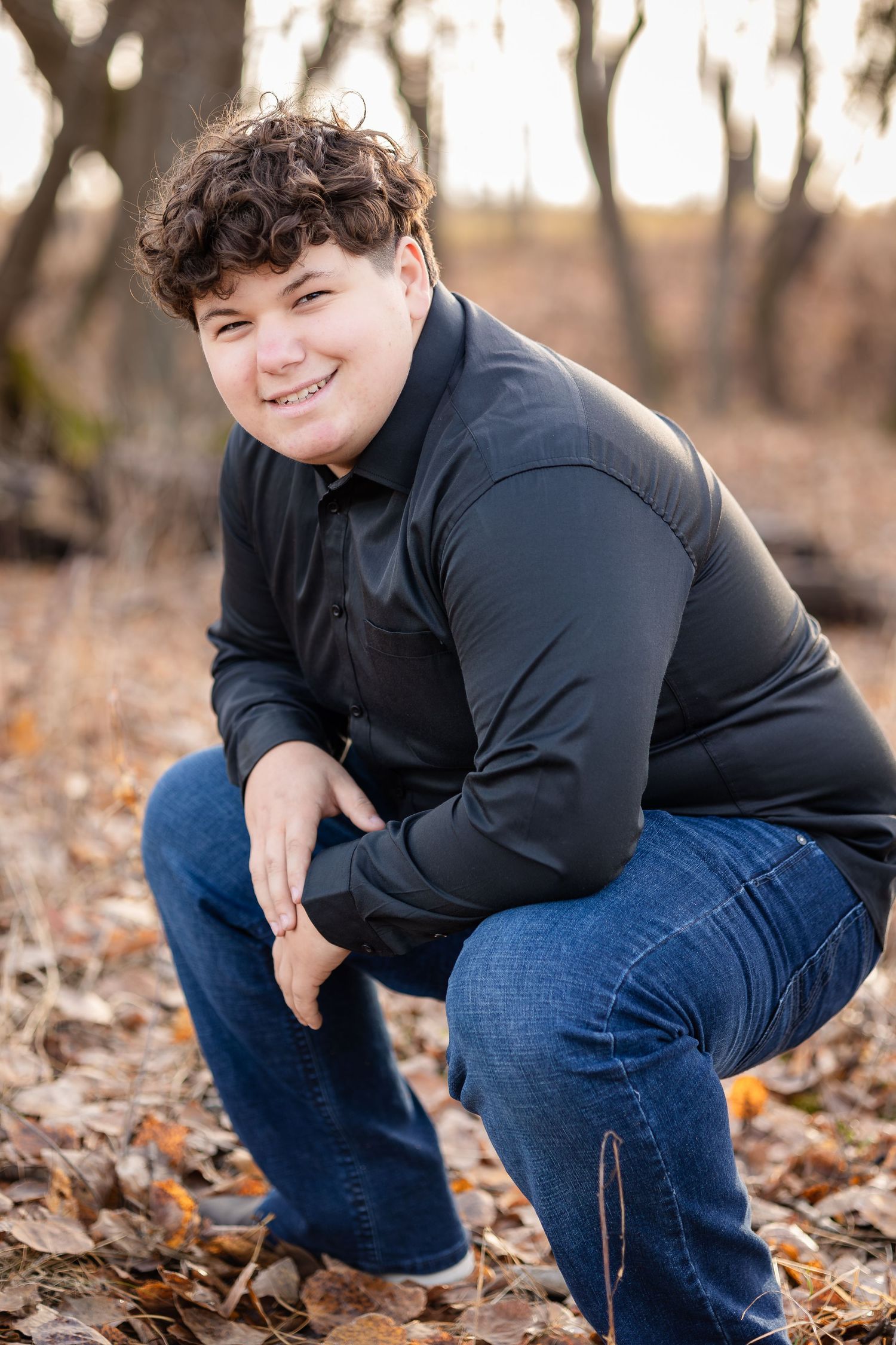 Person in black sweater crouching among fallen autumn leaves in wooded setting.