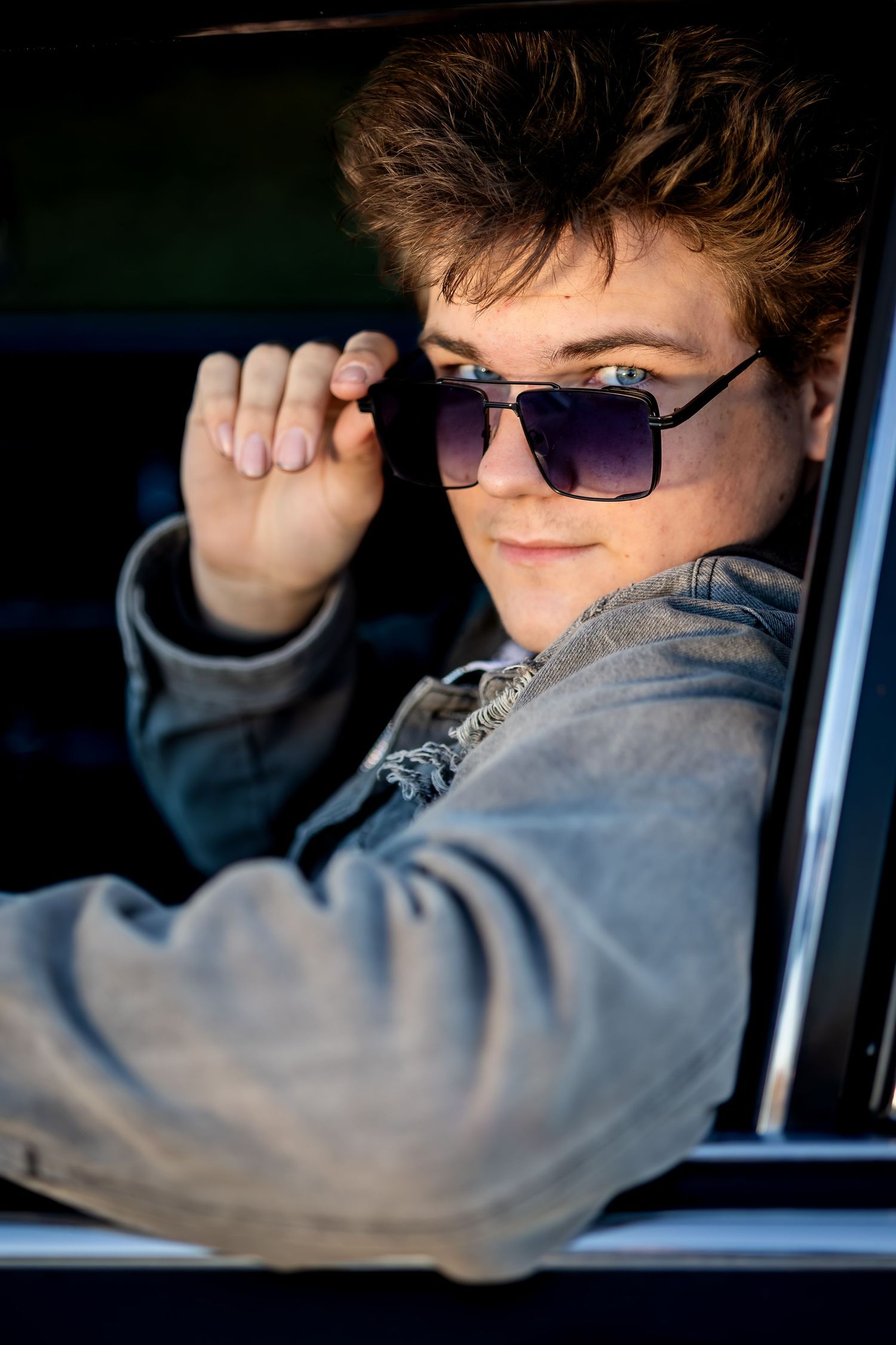 A person wearing sunglasses and a denim jacket leans out of a car window in dramatic lighting.