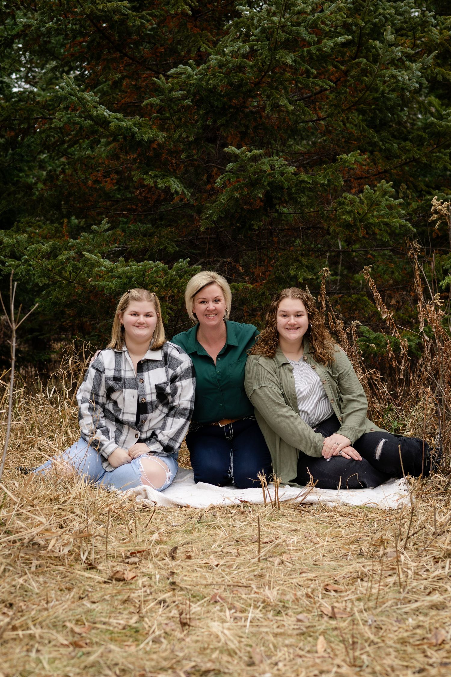Family group relaxes on the ground in an outdoor setting with trees and tall grass in the background.