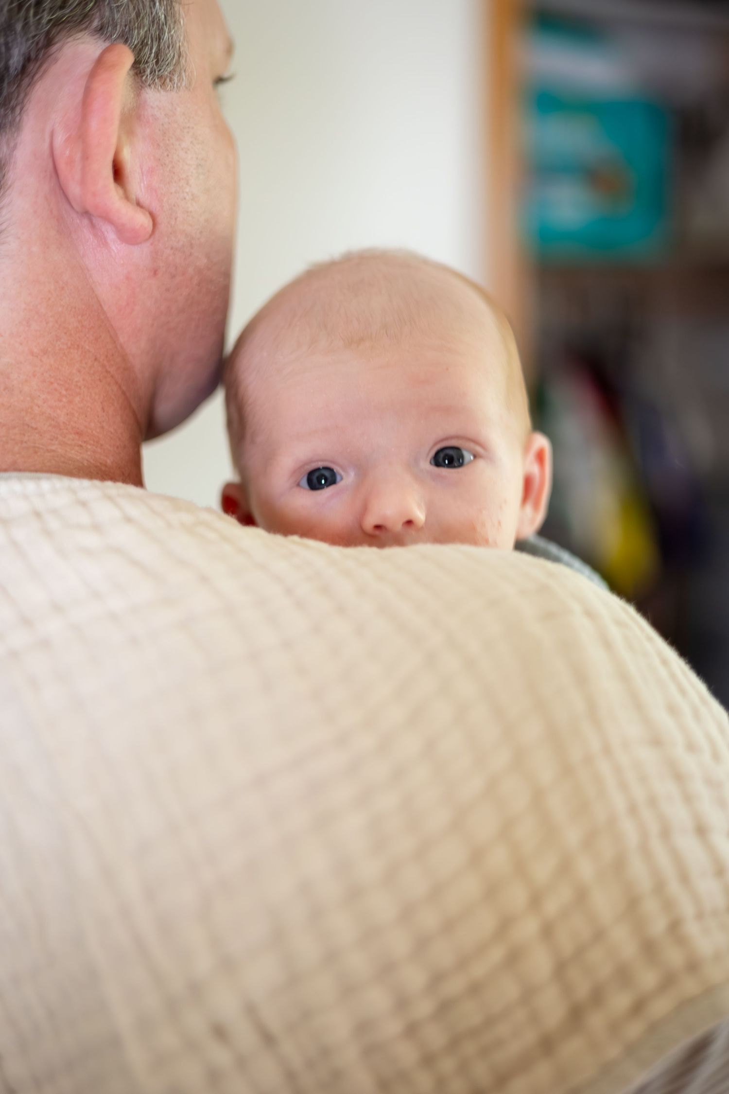 Baby peering over parent's shoulder in soft natural lighting.