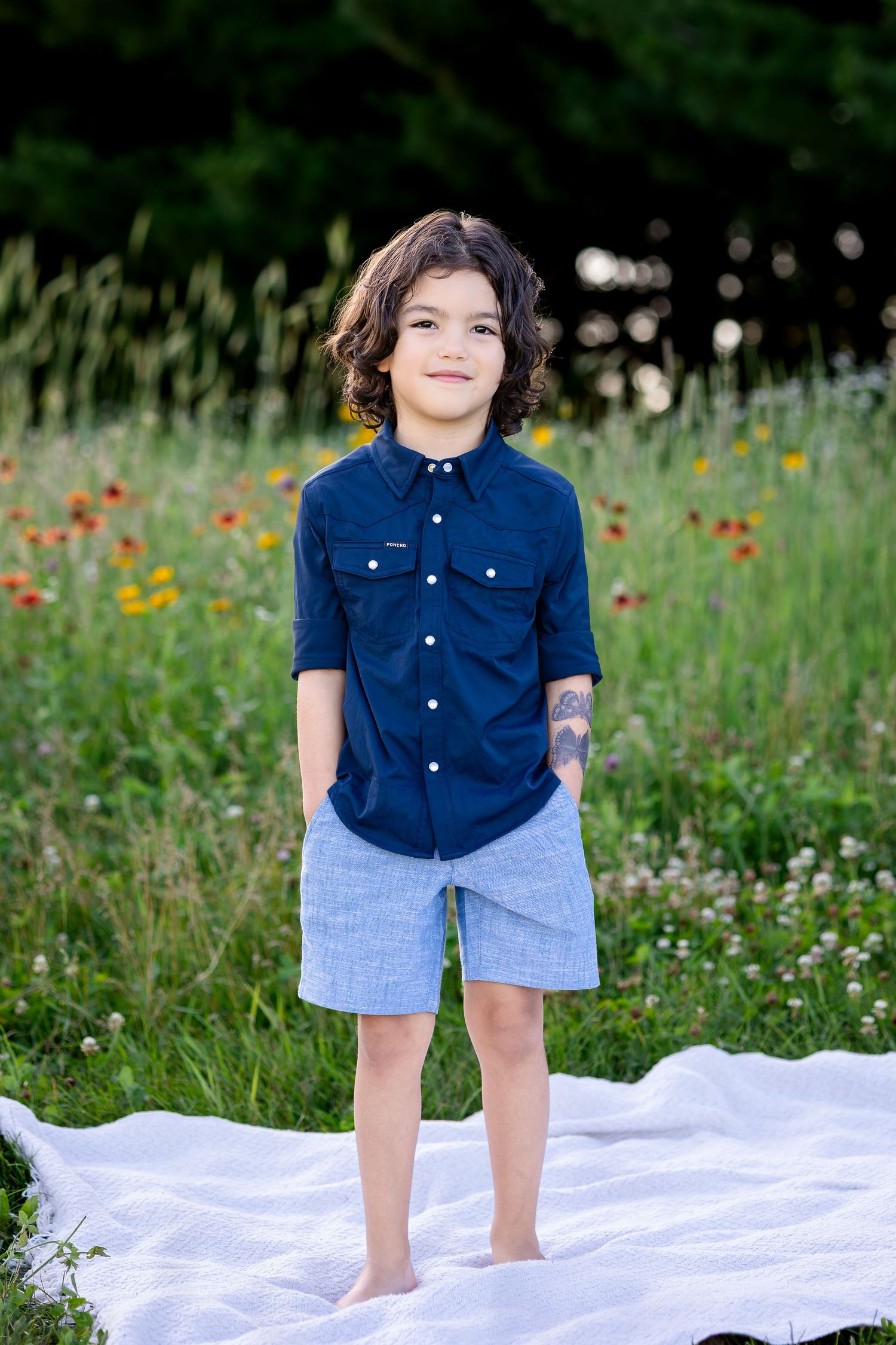A child in a navy blue shirt and denim shorts stands on a white blanket in a wildflower meadow with orange blooms.