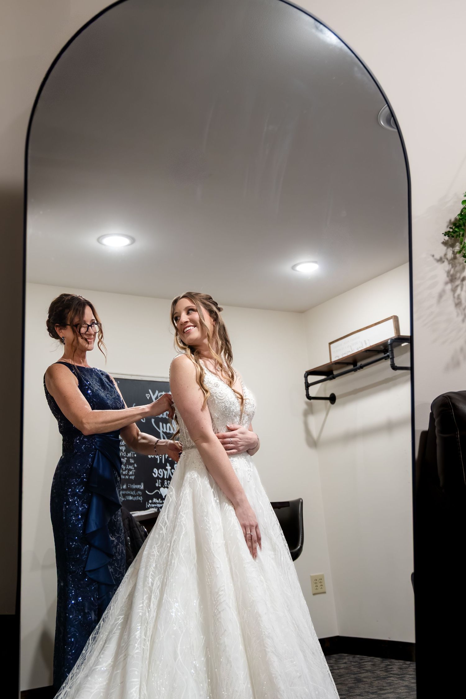 Bride smiling in white lace ball gown while woman in navy sequin dress helps with dress in mirror.