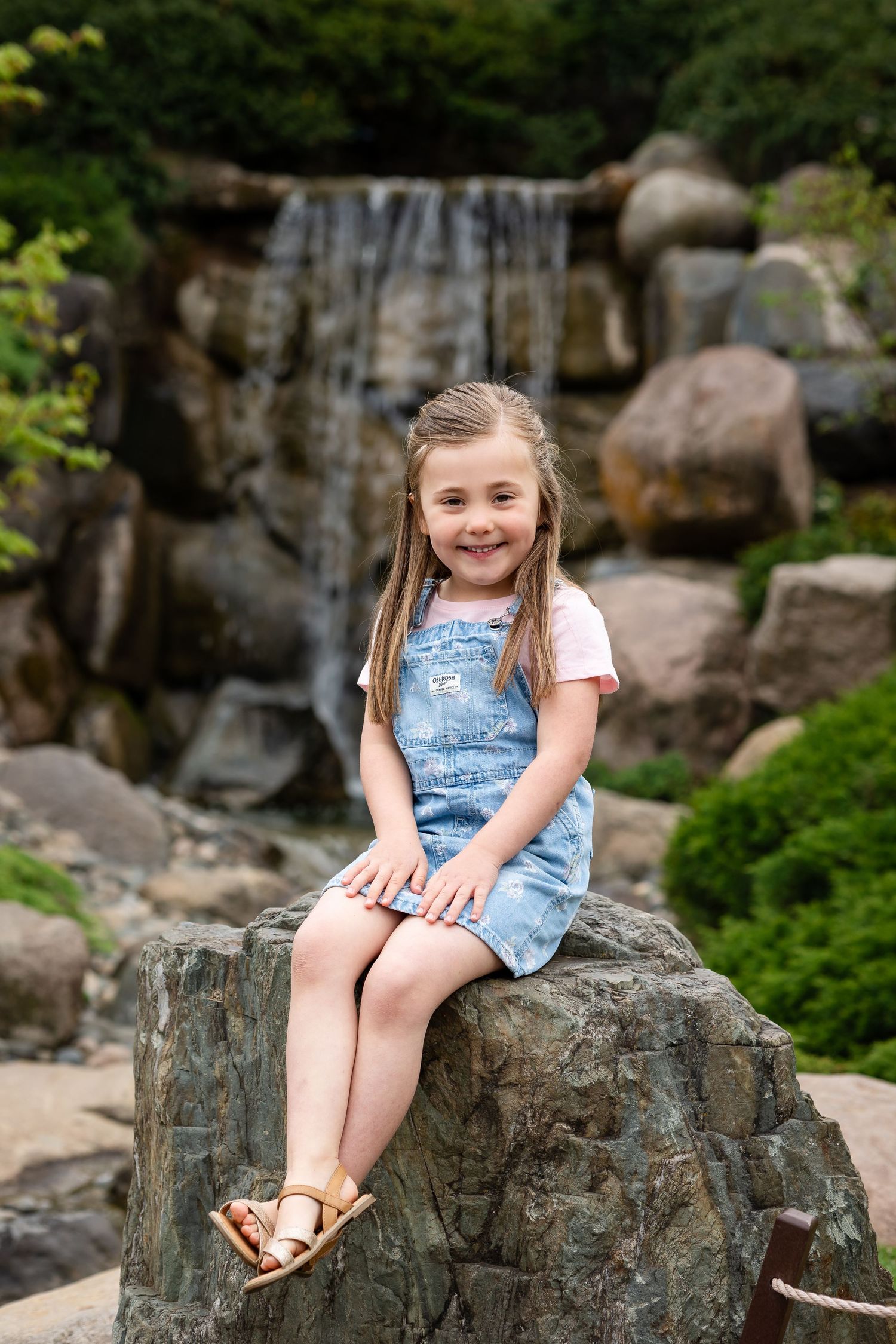 Young girl in denim overalls sits smiling on a rock with a waterfall and lush greenery in the background.