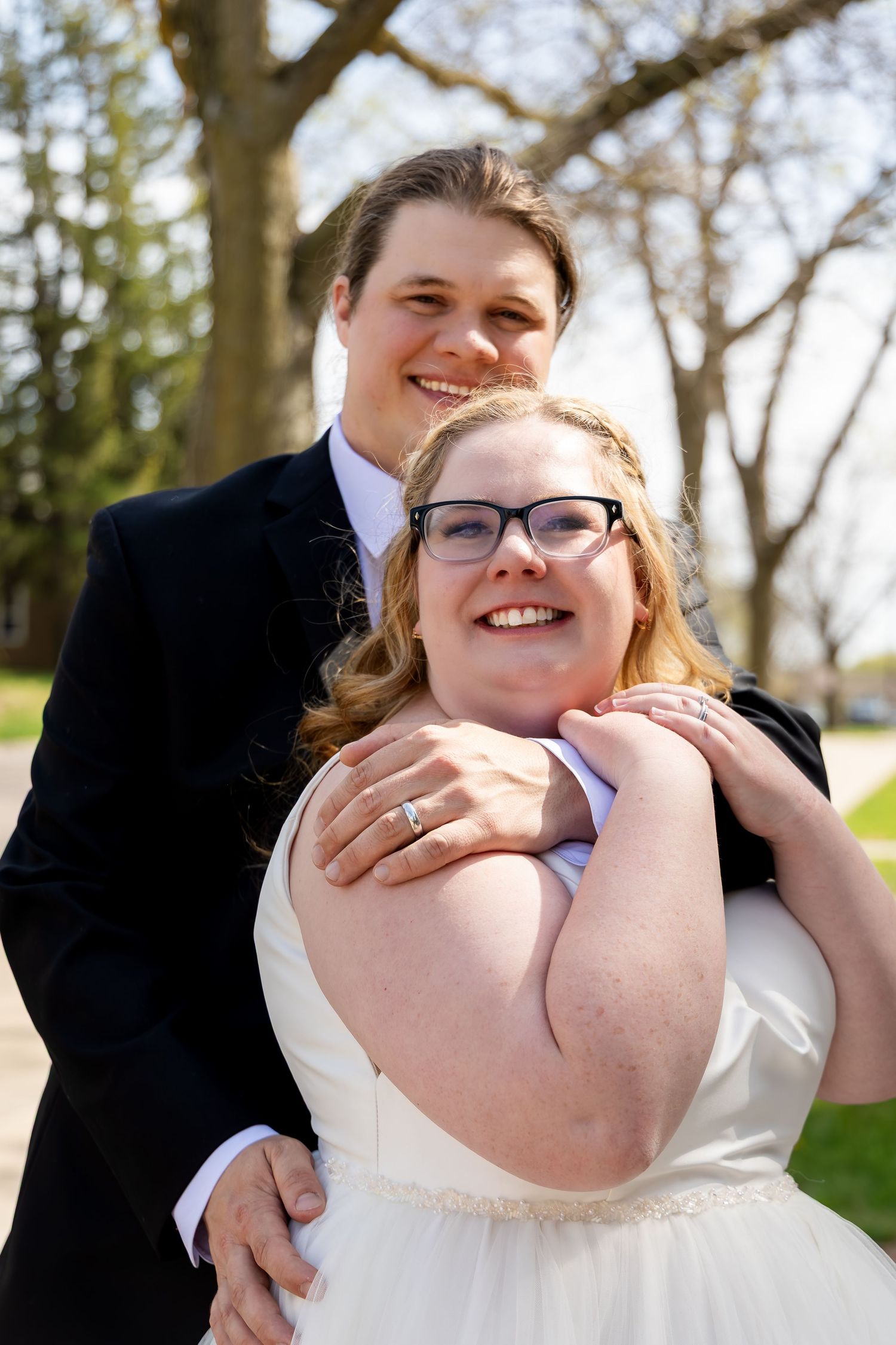 Groom in black suit embraces bride wearing white gown and glasses outdoors among spring trees.