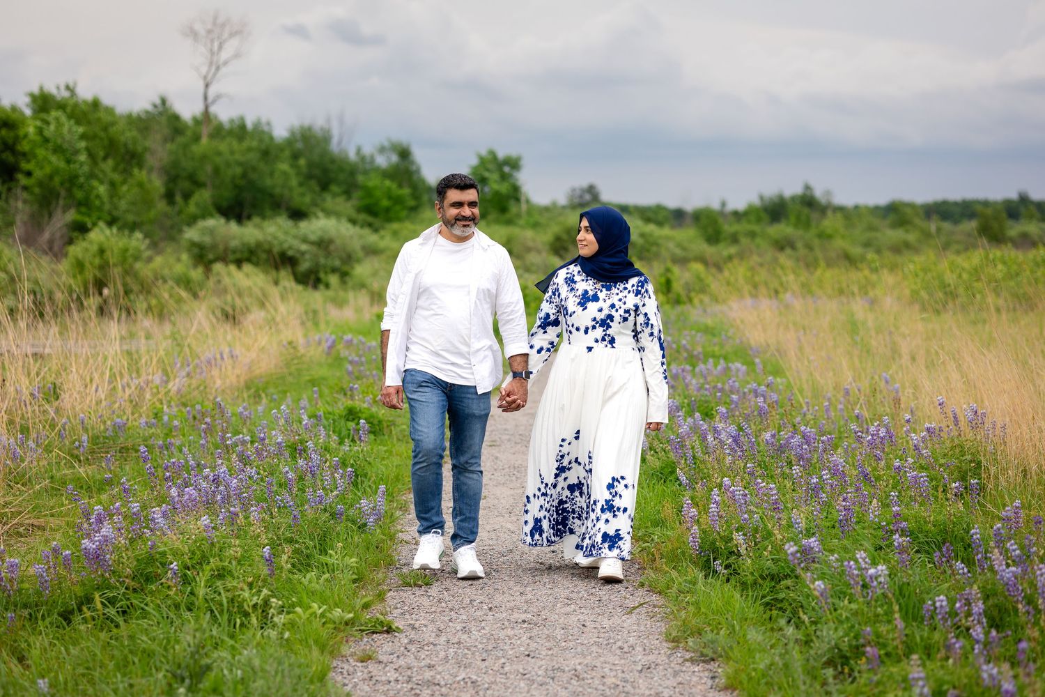 Couple walking hand in hand on a garden path surrounded by purple wildflowers and lush greenery.