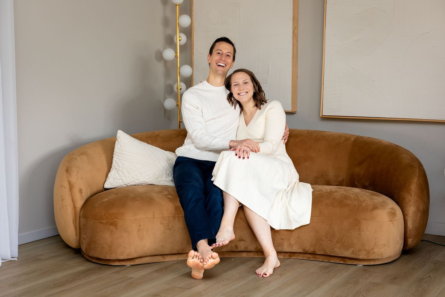 Couple smiling and embracing on a curved camel velvet sofa in a bright modern living room.