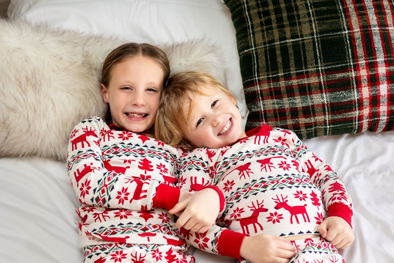Two children wearing matching red and white Christmas reindeer pajamas hugging and smiling on a white bed.