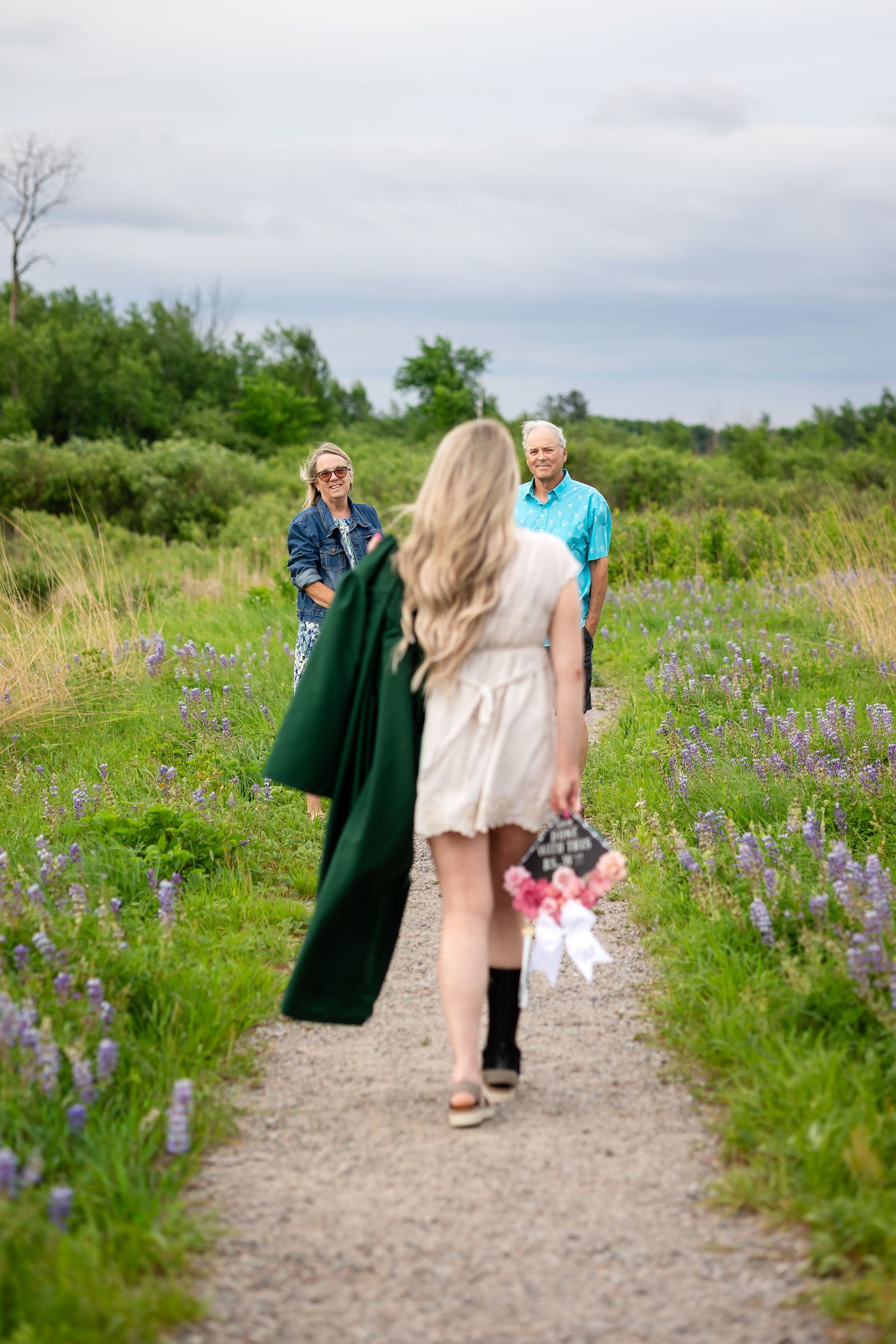 Blonde woman in white dress carrying graduation cap and bouquet walks toward elderly couple on a wildflower meadow path.