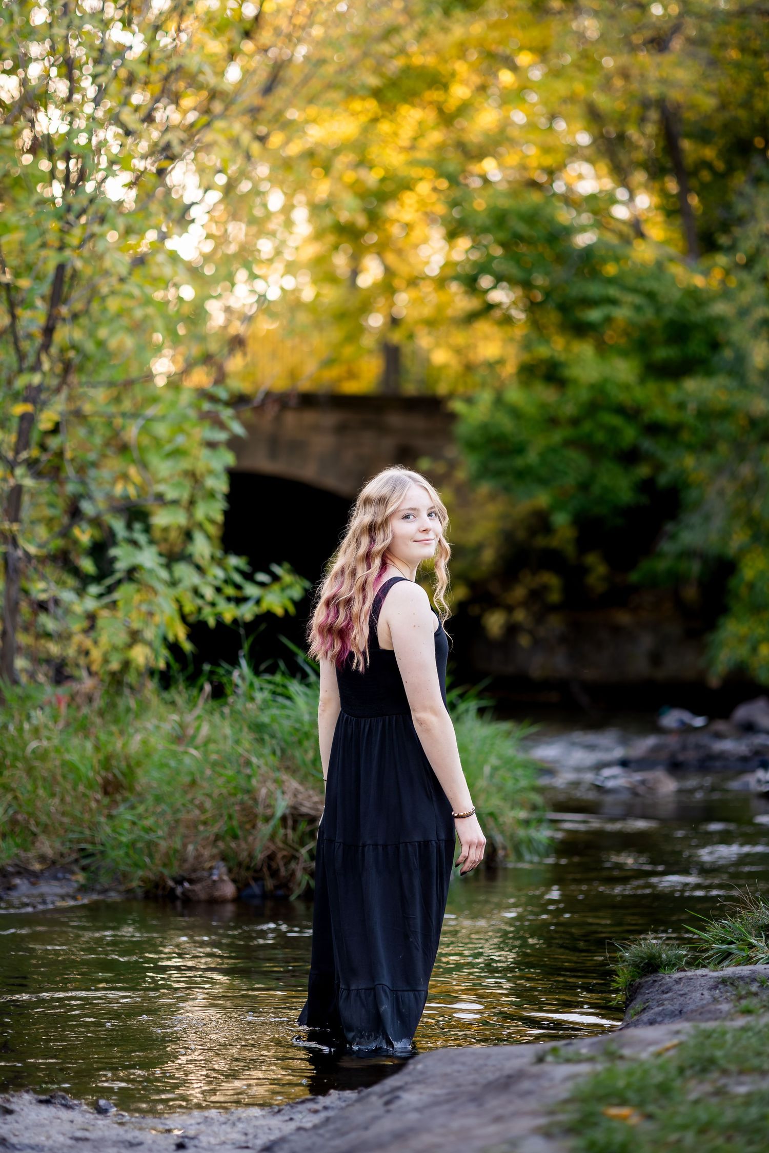 A person in a black dress sits by a tranquil stream surrounded by autumn foliage and golden sunlight.