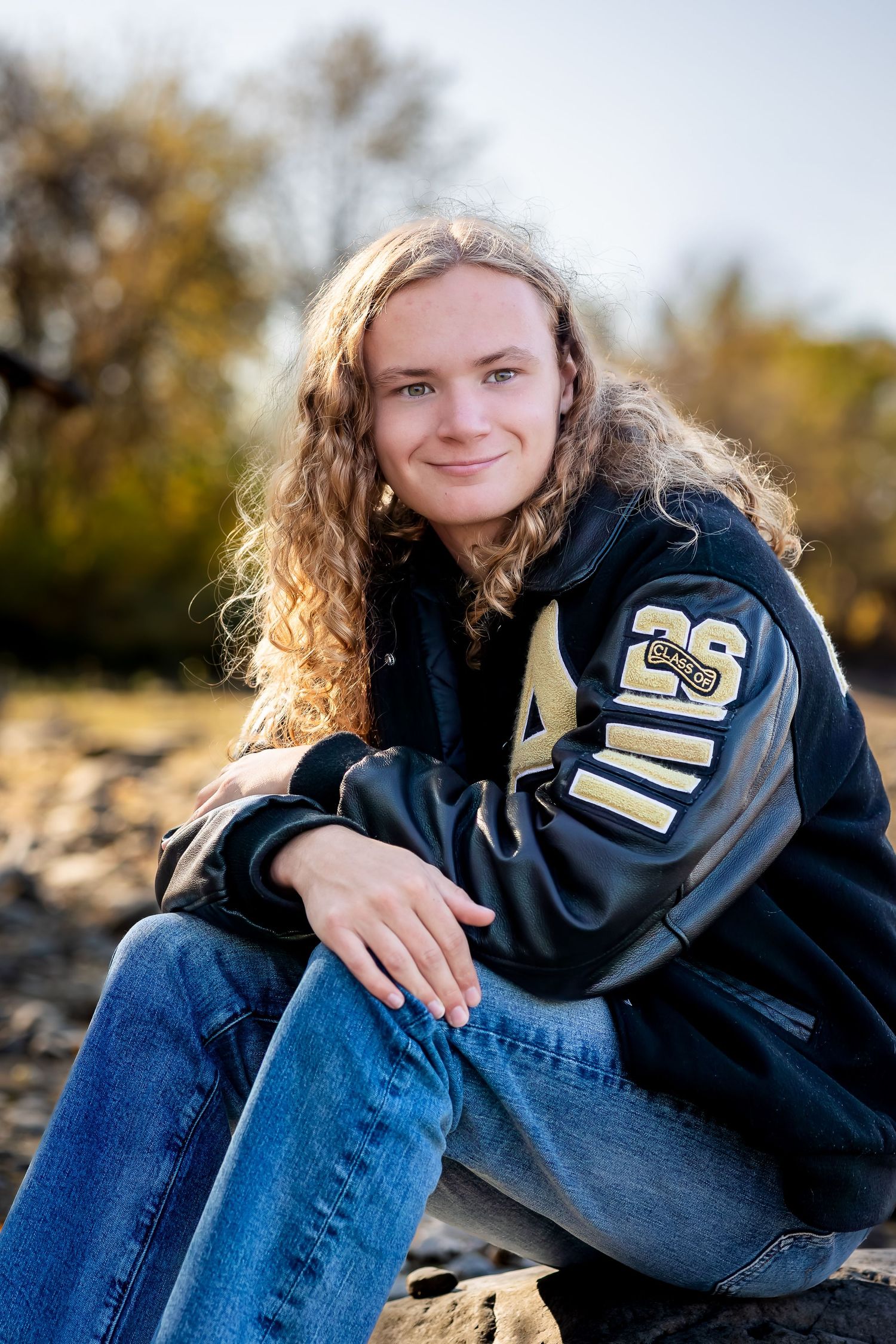 Student in a varsity letterman jacket sits casually on rocks during golden hour in an autumn setting.