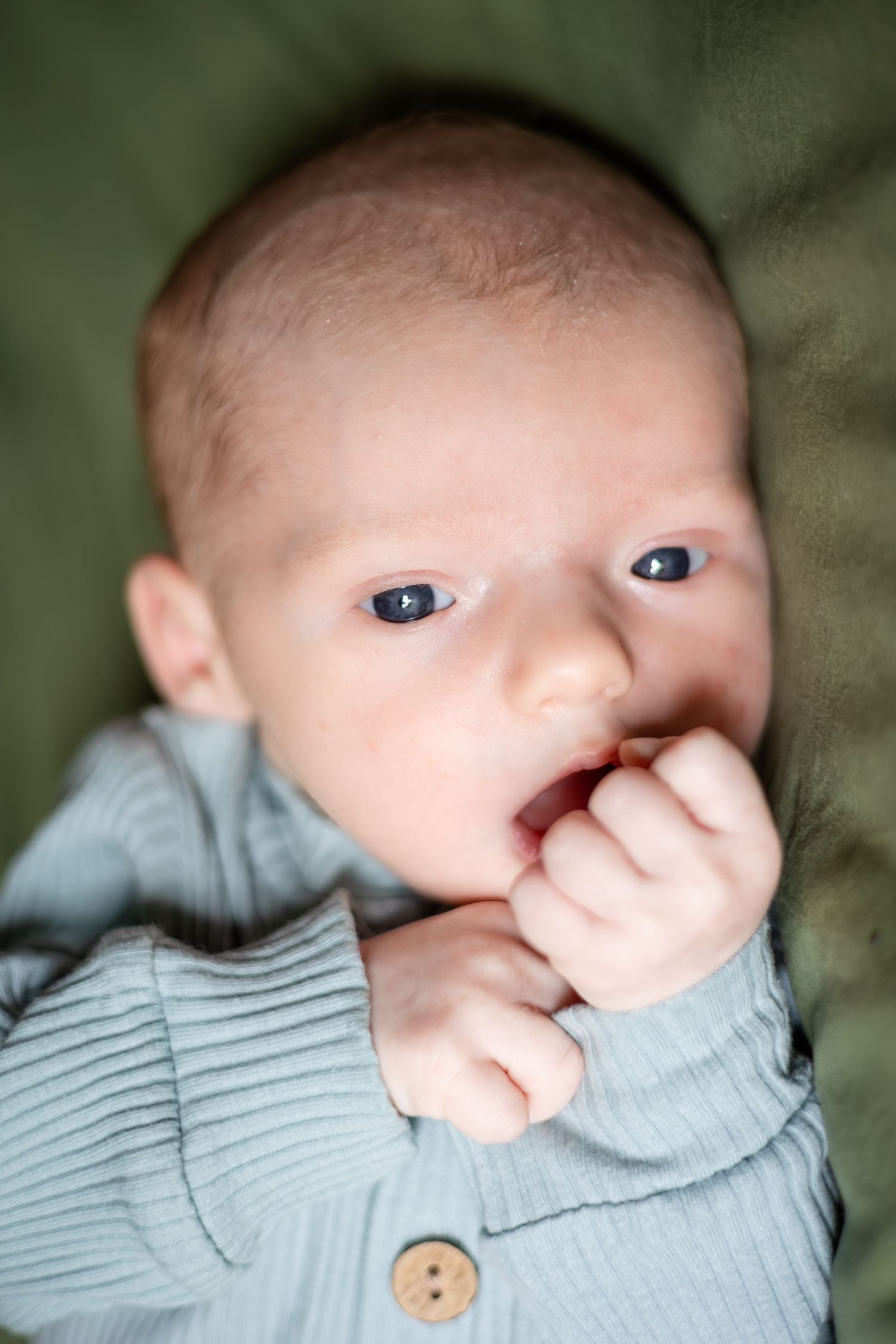Baby in blue ribbed sweater with hand near mouth lying on green background.