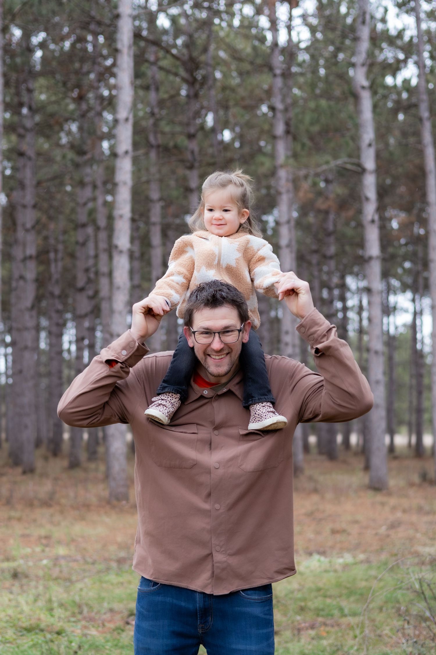 A playful moment captured as a toddler rides on shoulders in a pine forest.