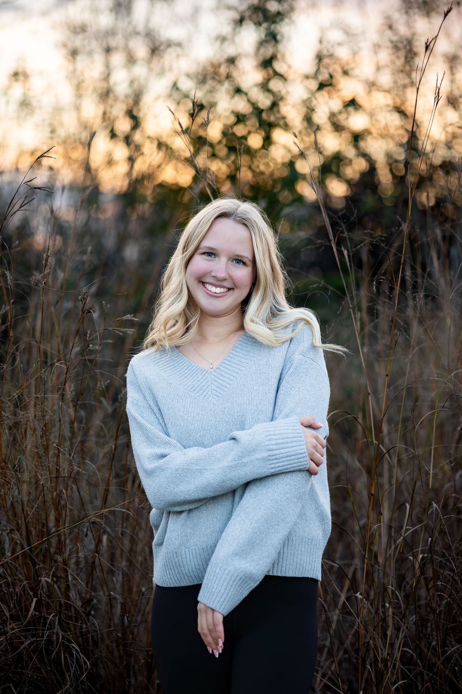 A person wearing a light gray sweater poses against tall dried grass and a sunset backdrop in a natural outdoor setting.