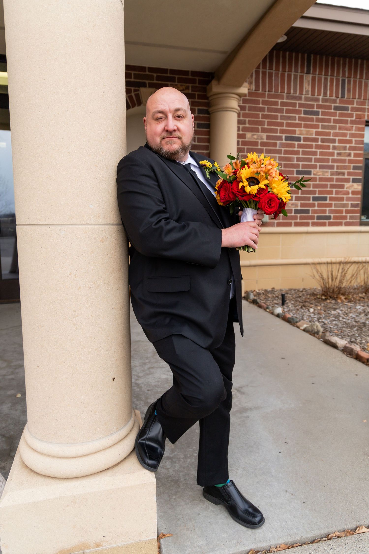 A person in a black suit poses with a vibrant sunflower bouquet against a brick building with columns.