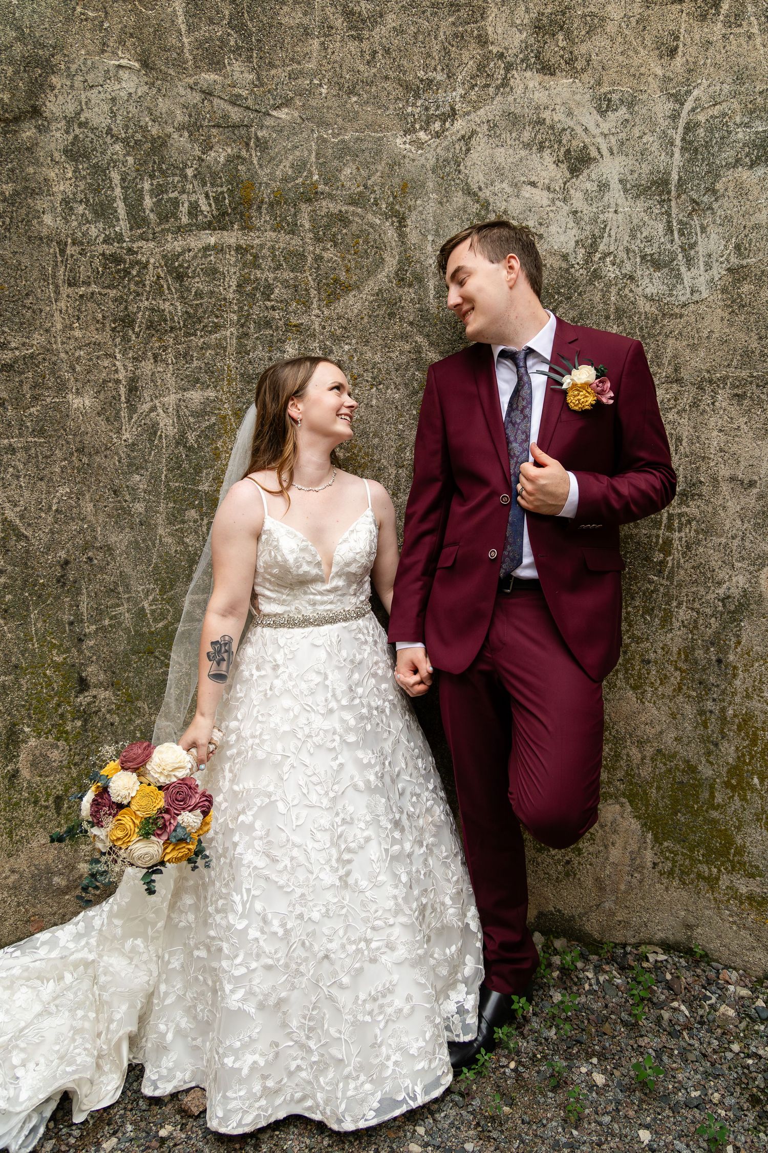 Wedding portraits of couple against textured wall with bride in white dress and groom in burgundy suit.
