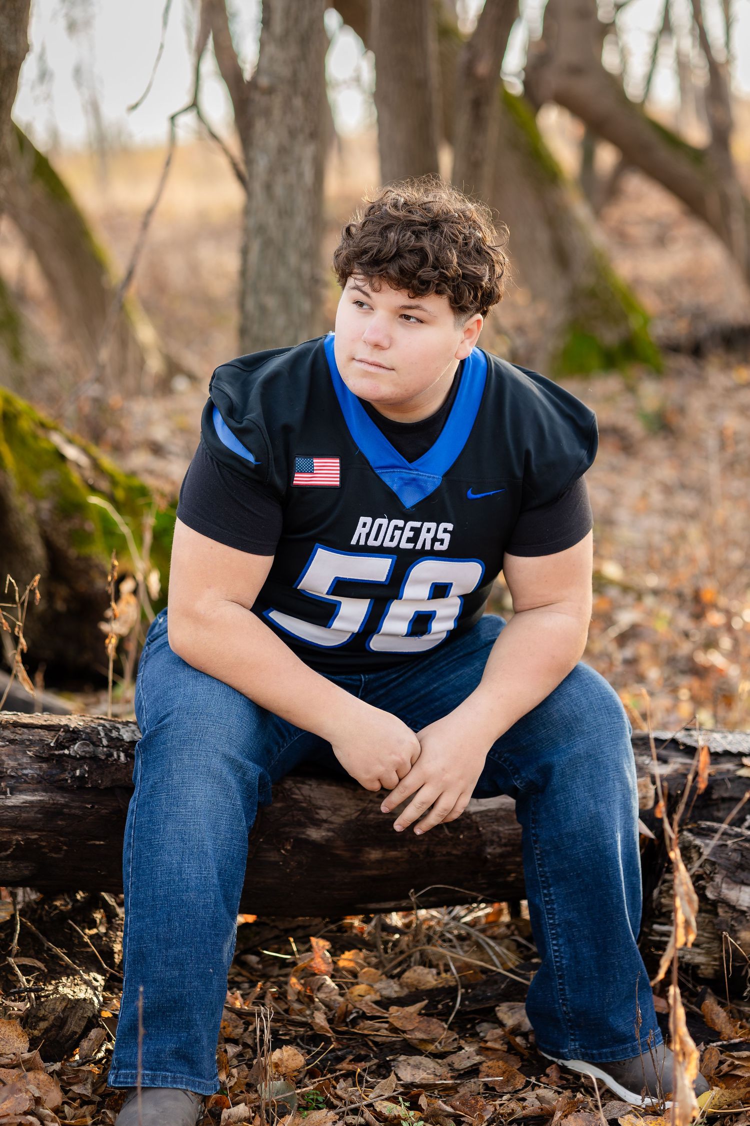 Person wearing a Rogers football jersey number 58 sitting on fallen logs in a wooded area.