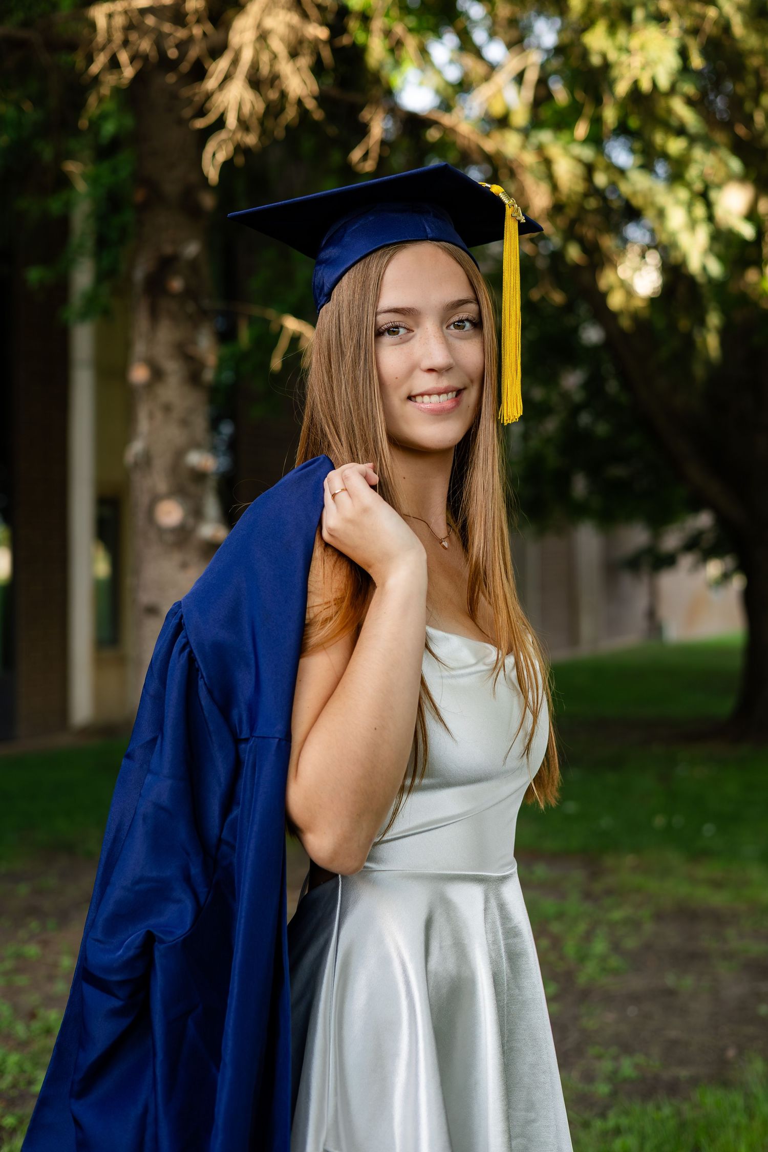 Graduate in blue cap and gown poses under trees during sunset.