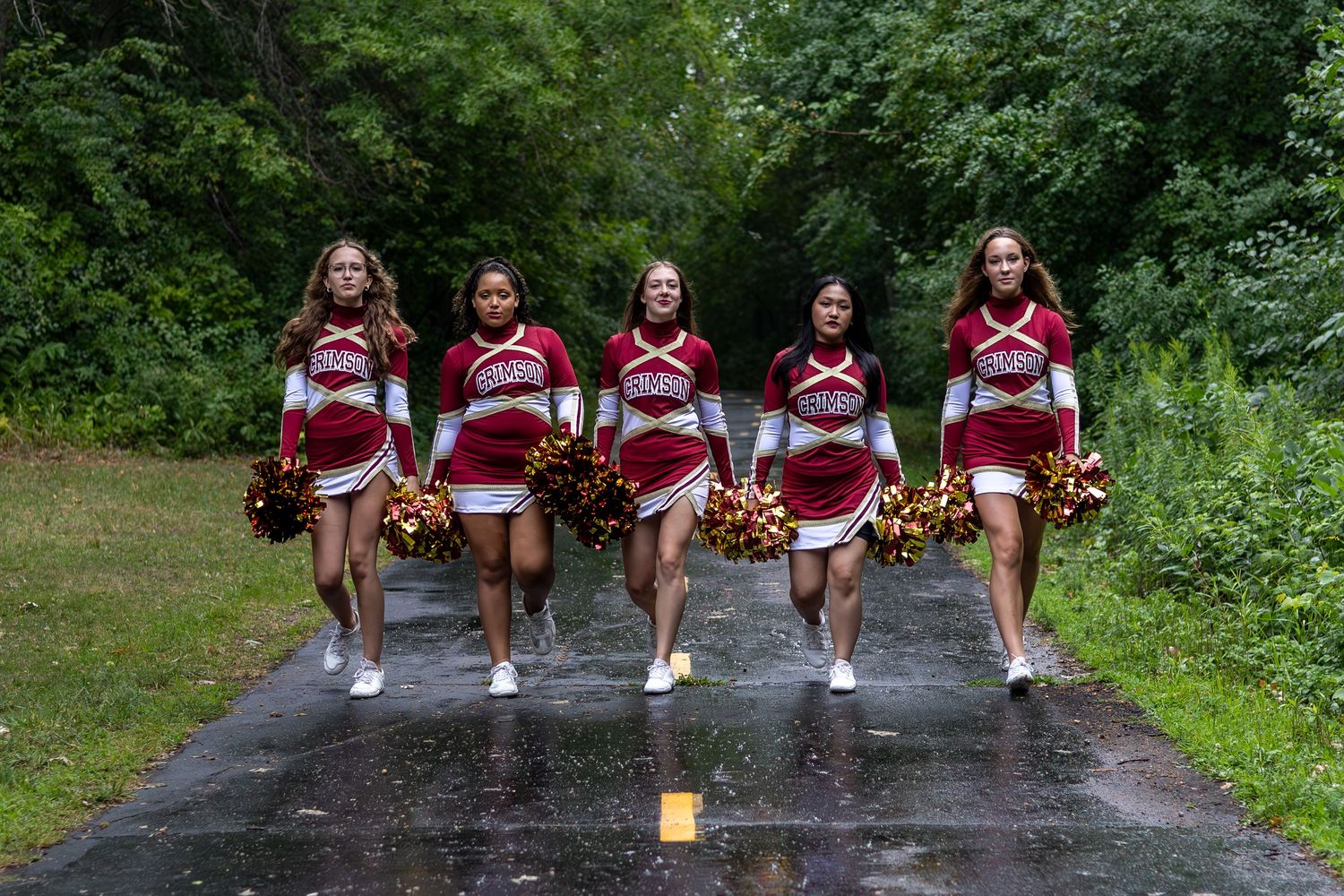 Five cheerleaders in red and white uniforms walking together on a path while holding pom poms.