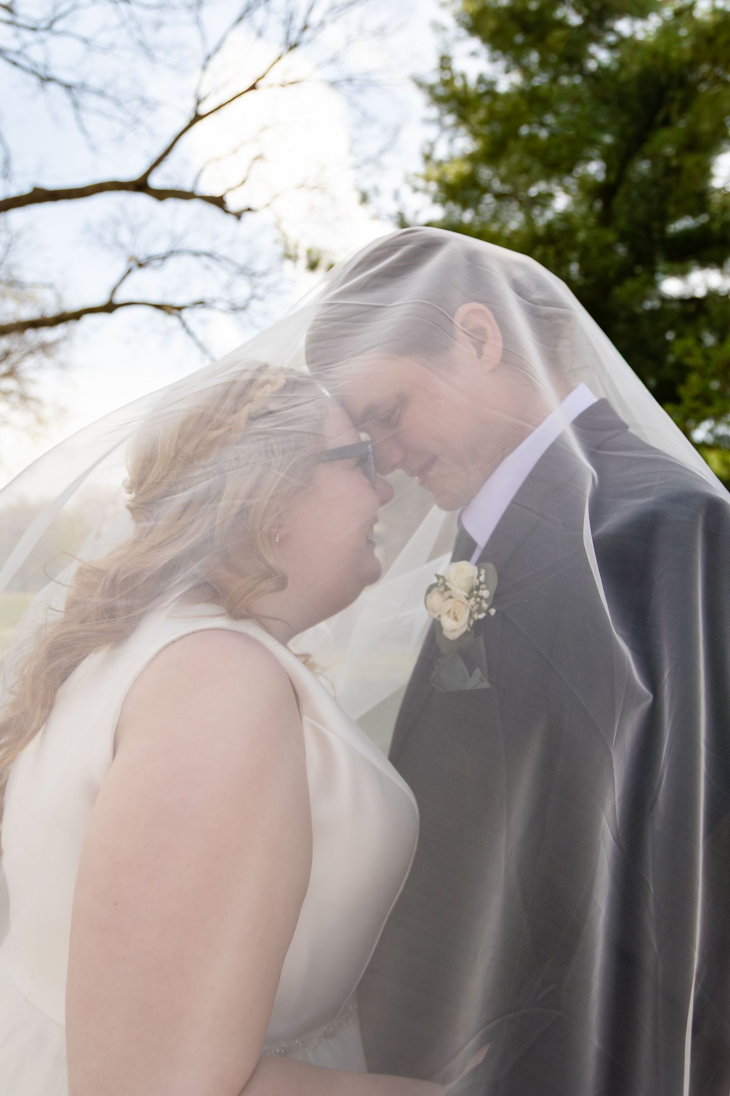 Bride and groom touch foreheads romantically under flowing white veil outdoors.