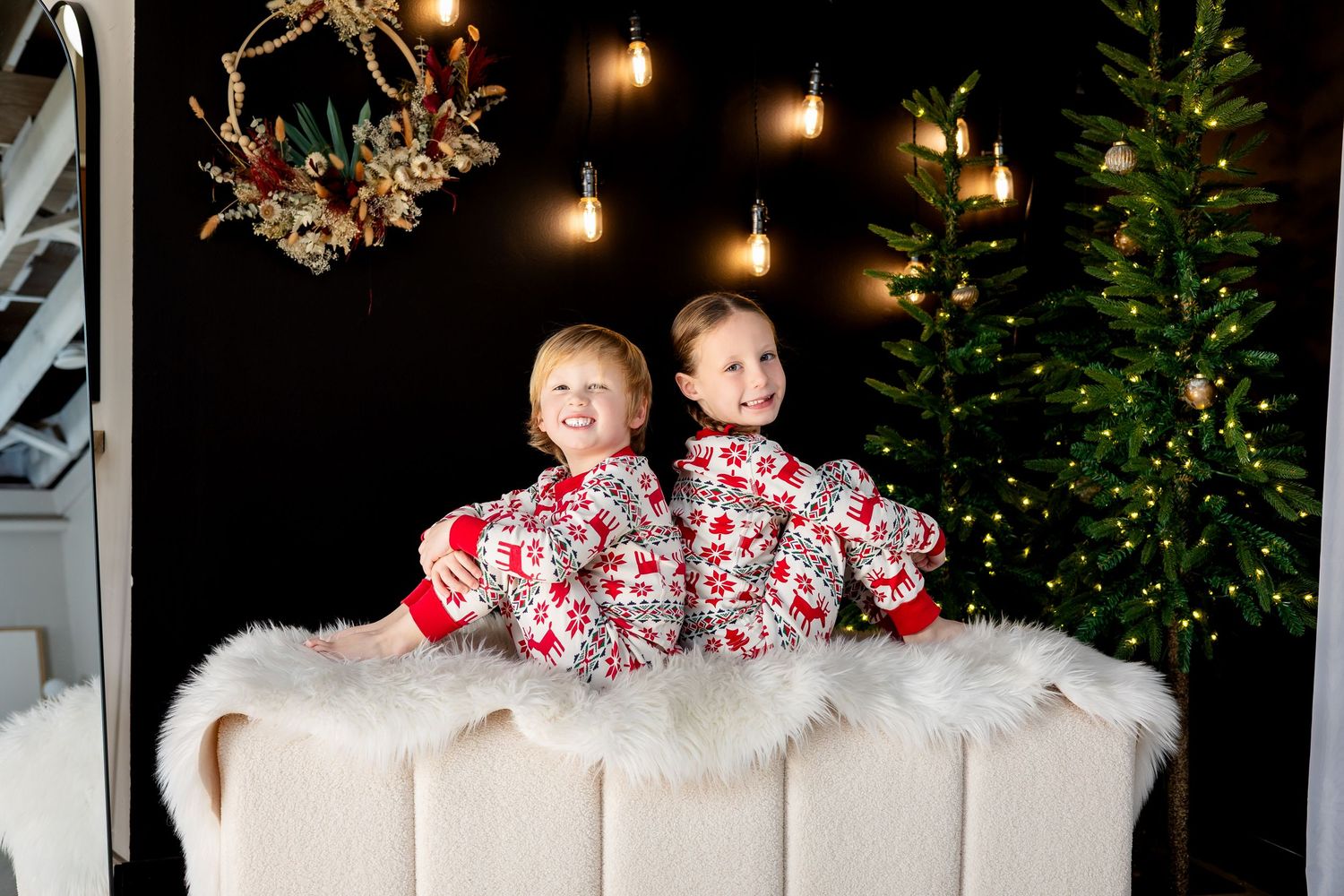 Two children in matching Christmas pajamas sit back-to-back on a white fur bench near a decorated holiday tree.
