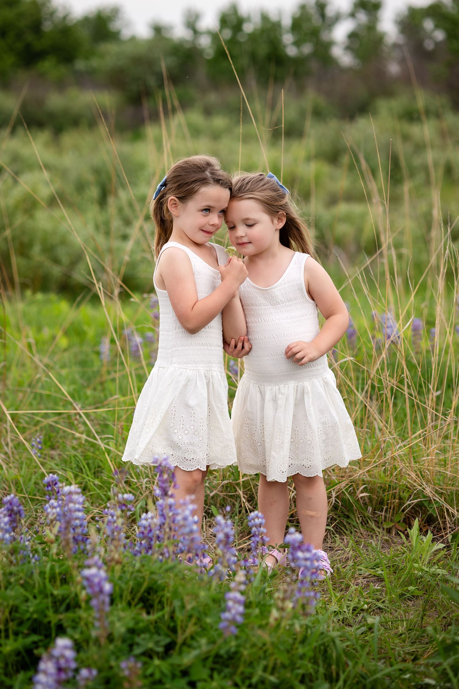 Two young girls in matching white dresses stand among purple wildflowers in a lush green field.