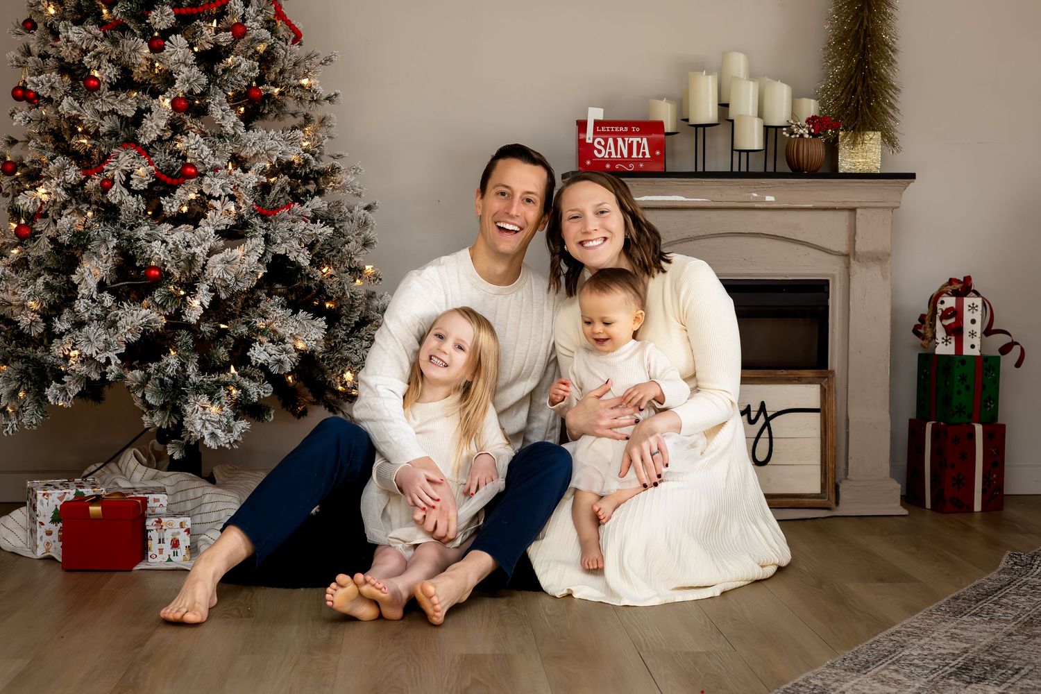 Family of four in matching white outfits smiling beside a decorated Christmas tree with gifts and fireplace.