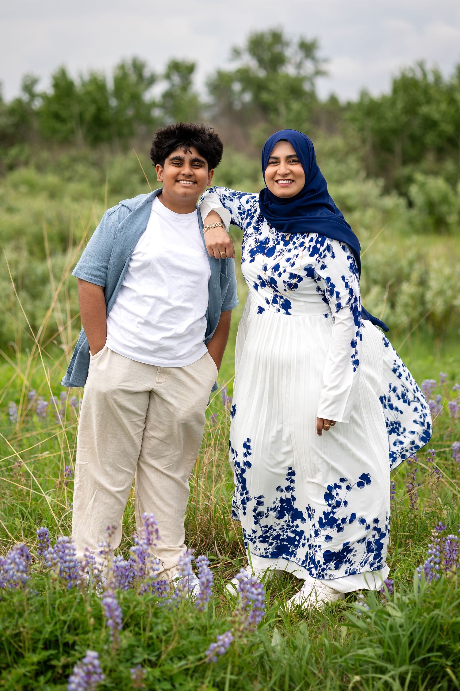 A young man and woman in a hijab and floral dress smile together in a wildflower field.