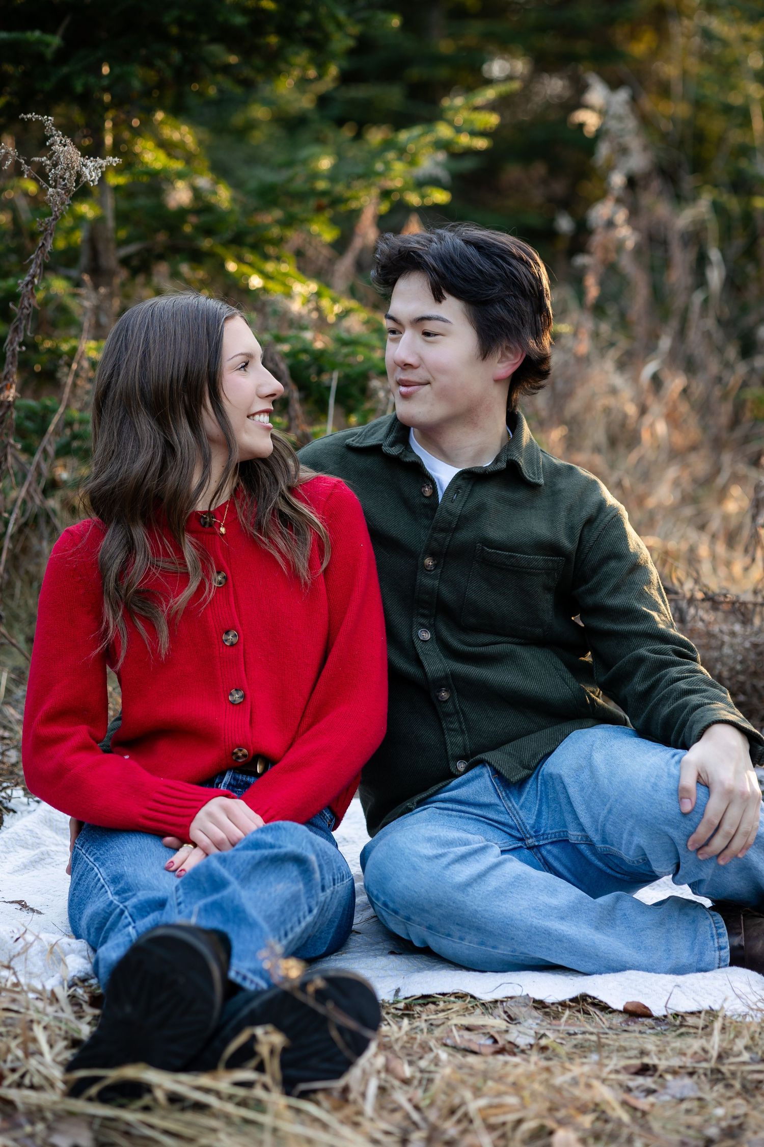A couple in red and dark jackets share a relaxed moment on fallen leaves.