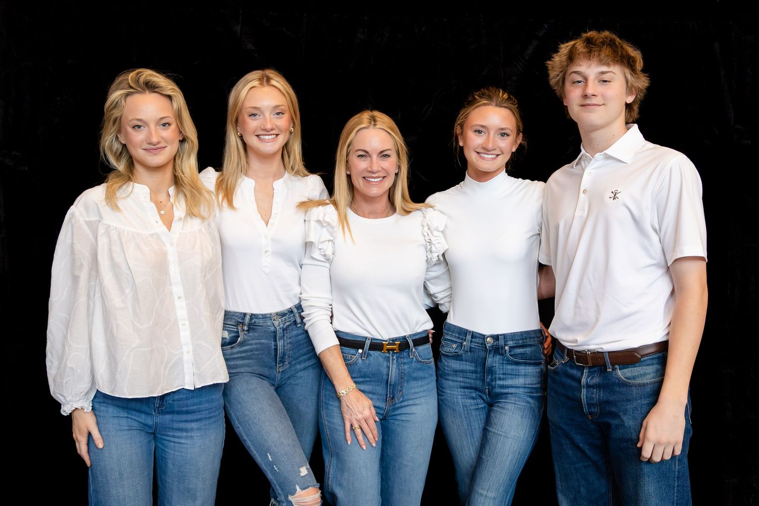 Group wearing coordinated white tops and blue jeans posing together against dark background.