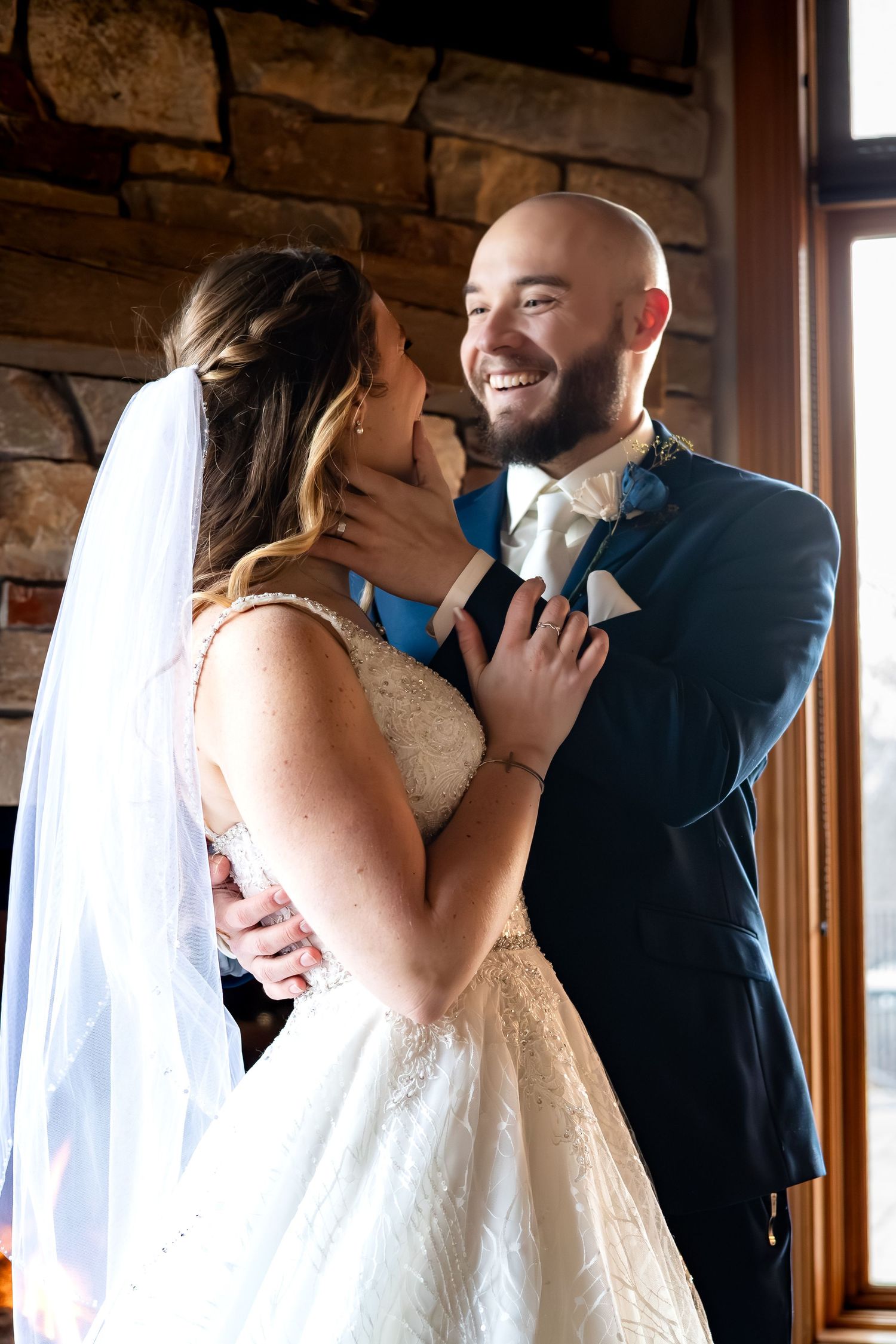 Bald groom in navy suit smiling joyfully at bride in lace gown during first look at rustic venue.