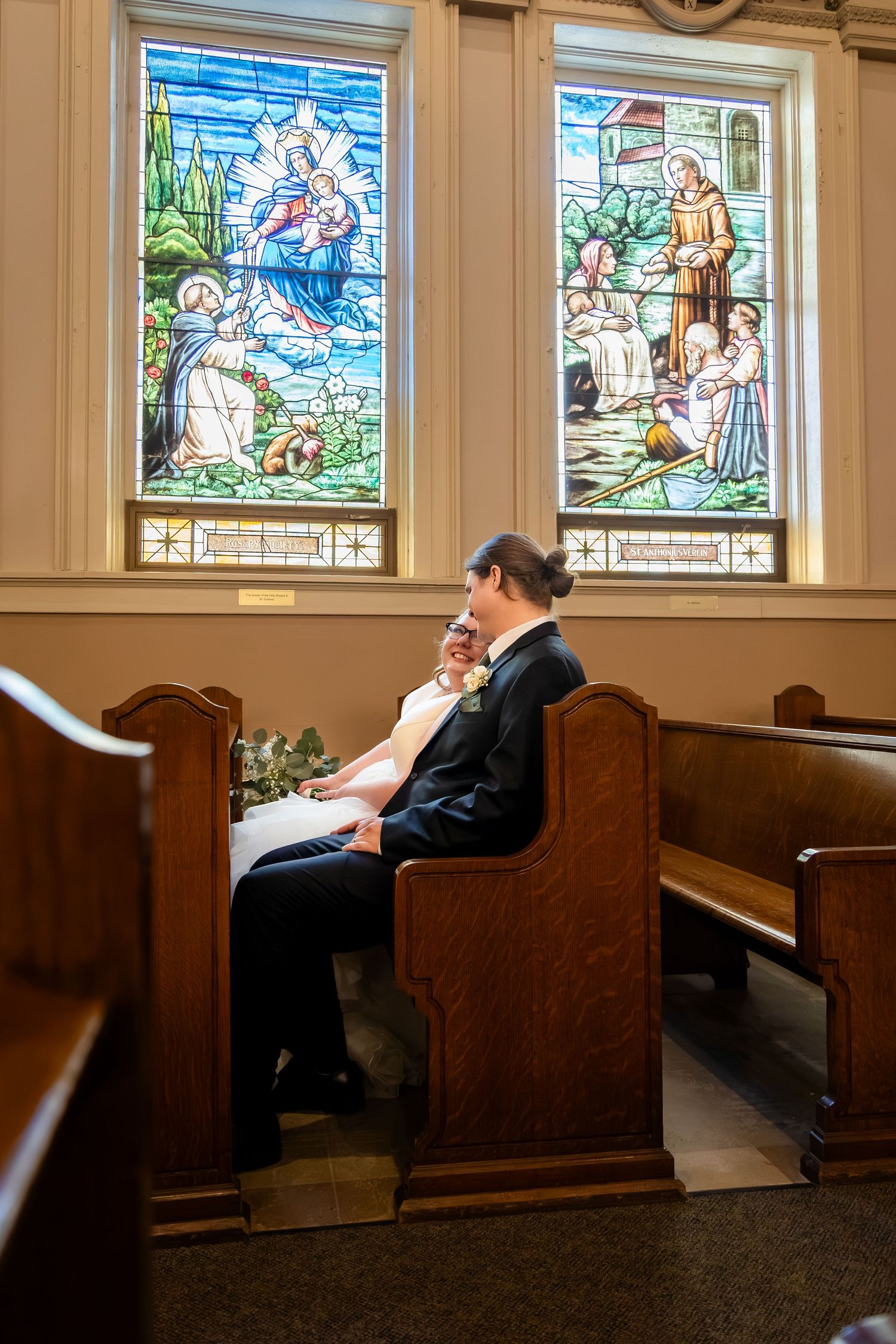 Bride and groom sitting intimately in church pew before stunning stained glass windows.