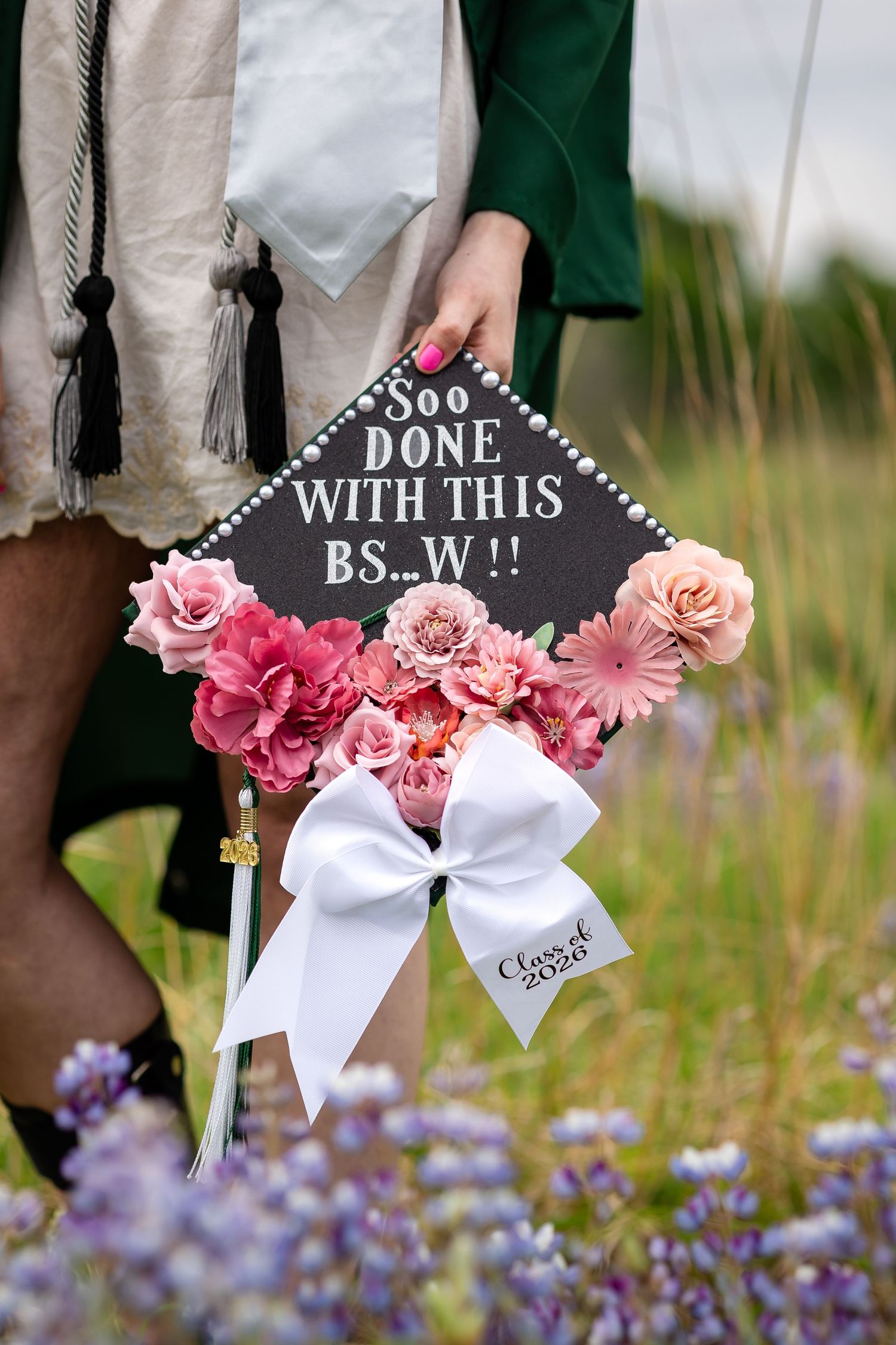 Decorated graduation cap with pink flowers, white bow, and humorous BSW degree quote, held outdoors among purple wildflowers.