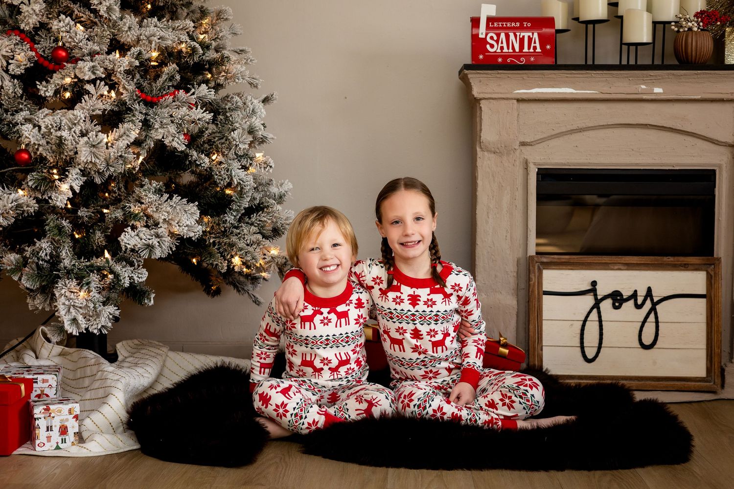 Two children in matching Christmas pajamas sit smiling by a decorated tree and fireplace with a Joy sign.