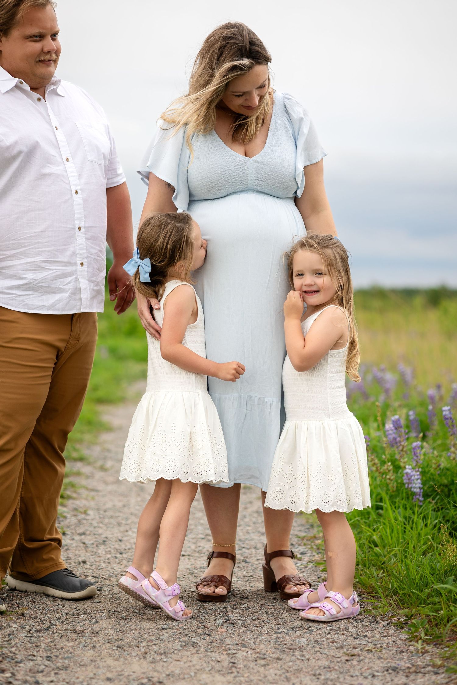 Two little girls in white dresses lovingly touch their mothers pregnant belly outdoors near lupine flowers.