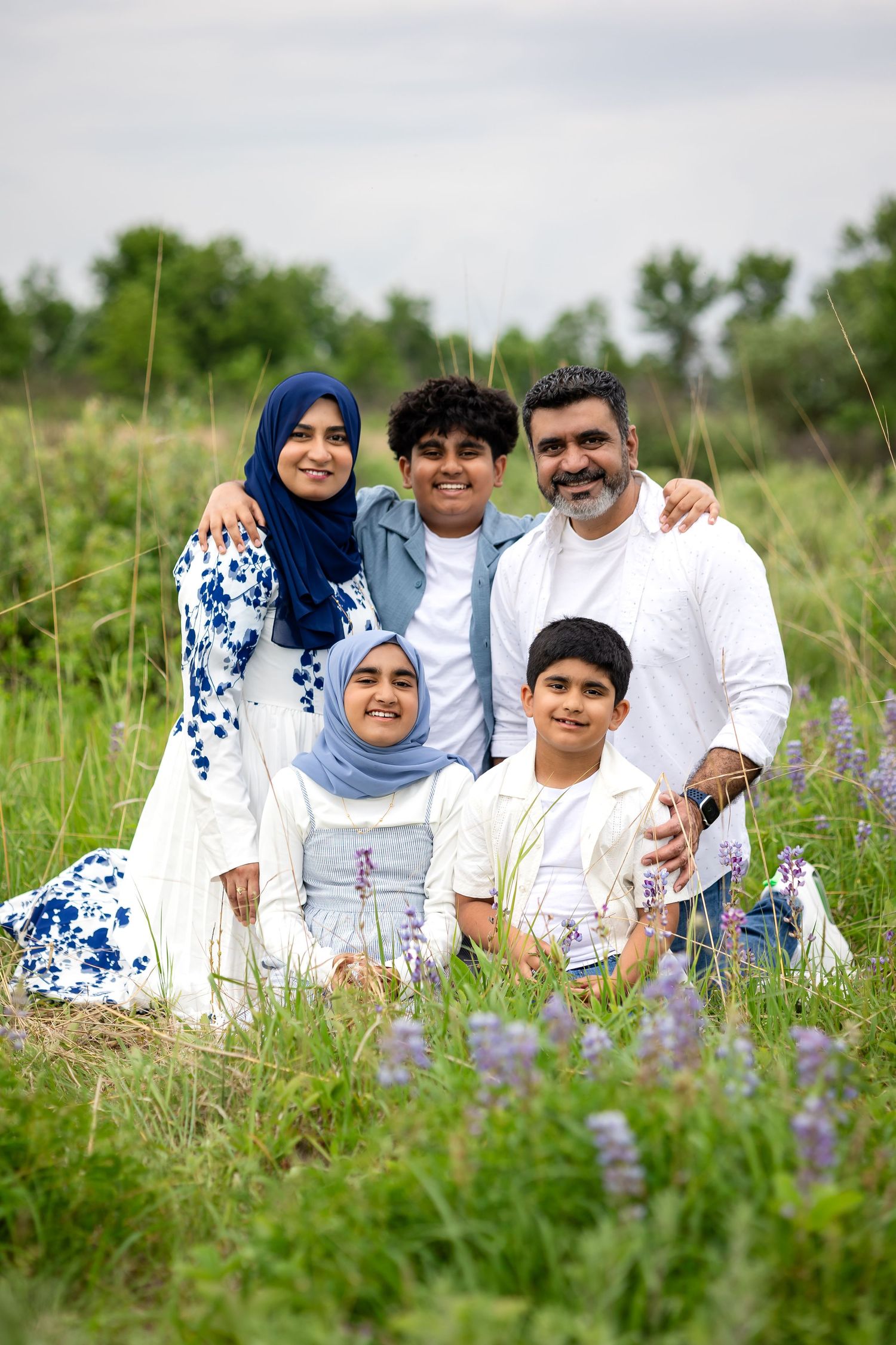 Muslim family of five posing together in a green field with purple wildflowers on a cloudy day.