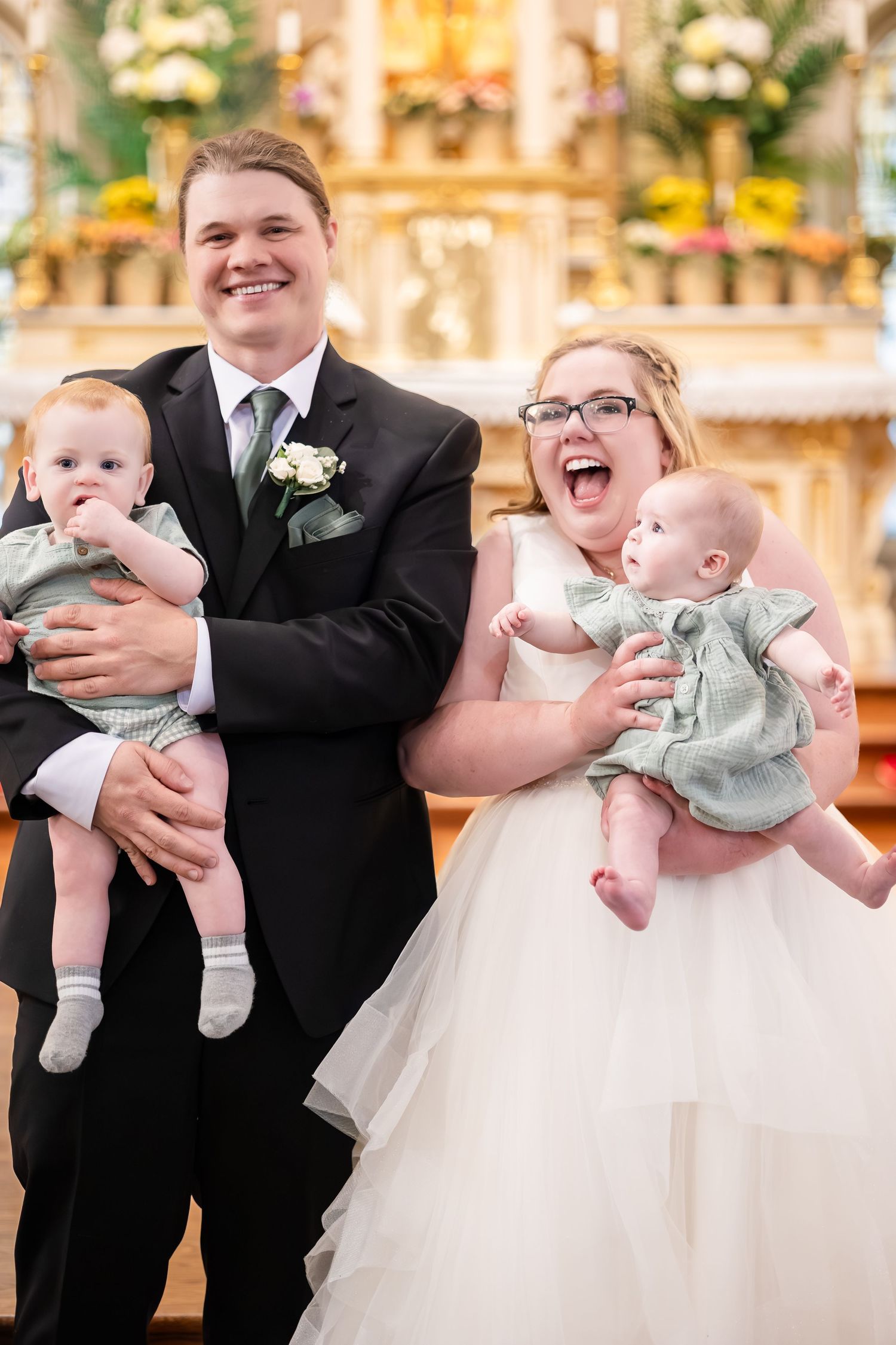 Laughing bride and groom in wedding attire each holding a baby in matching green outfits at church altar.