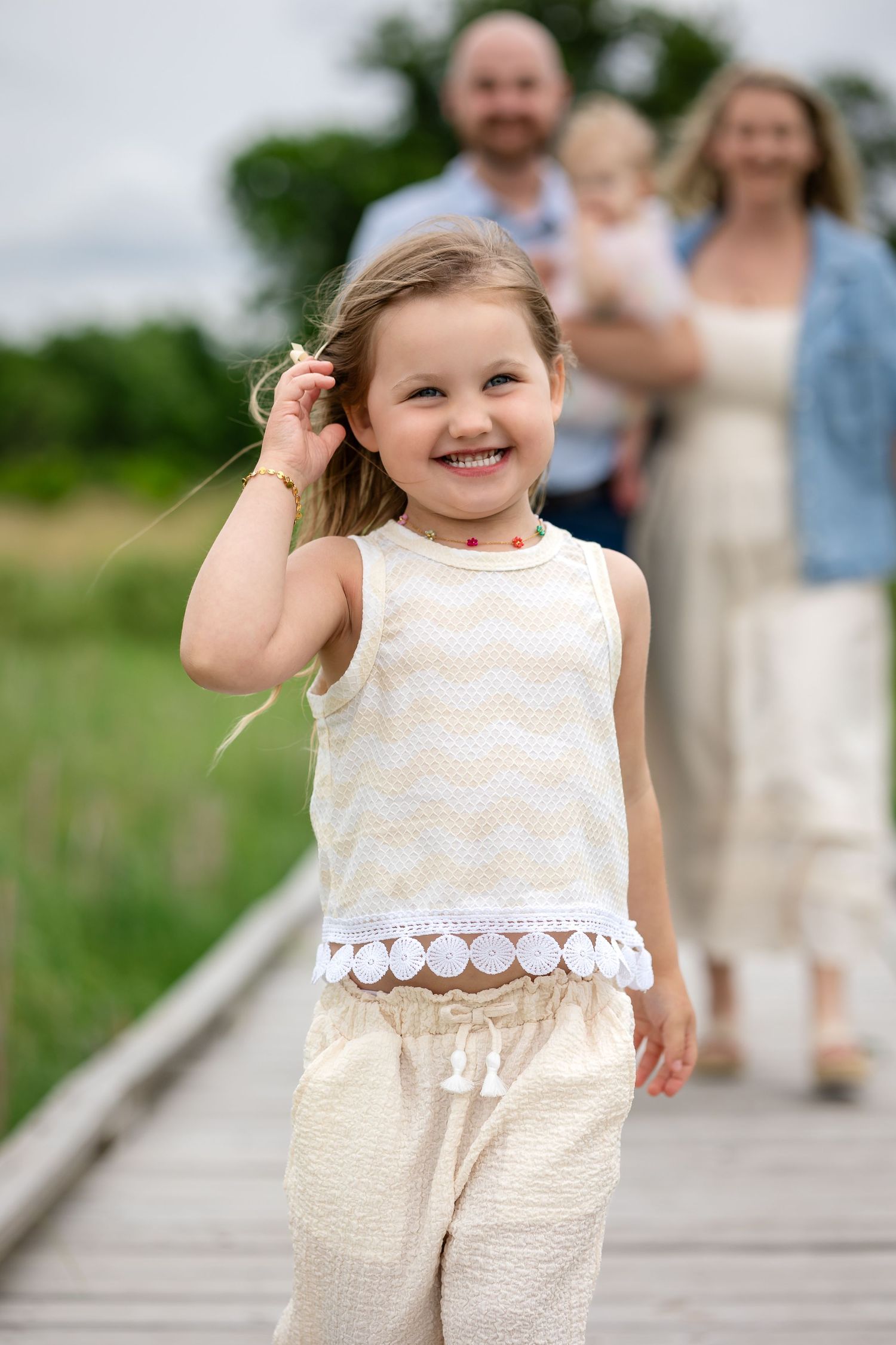 Happy little girl in white outfit laughing on wooden boardwalk with family walking behind her.