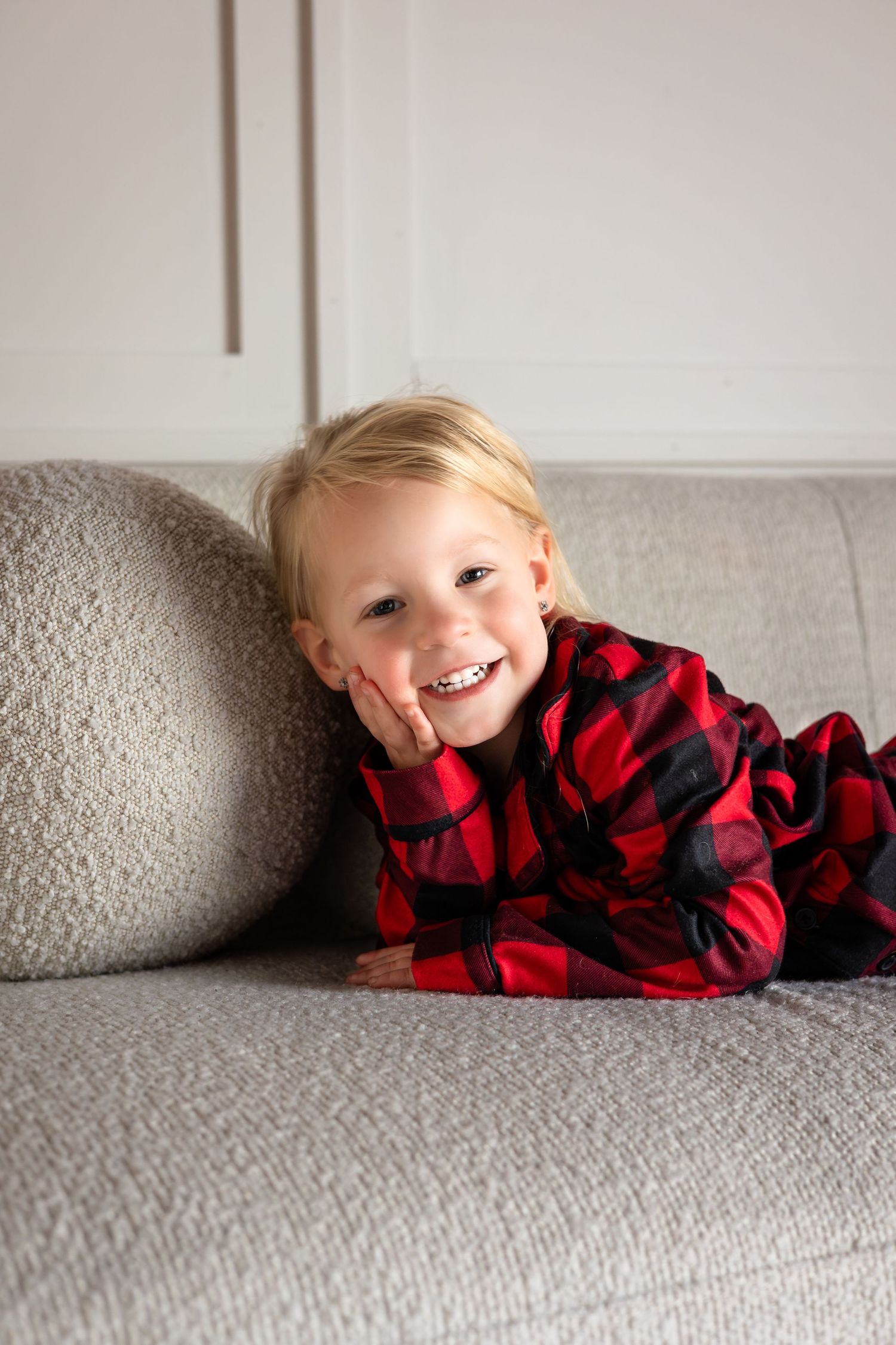 Child in red buffalo plaid pajamas lying playfully on gray couch.