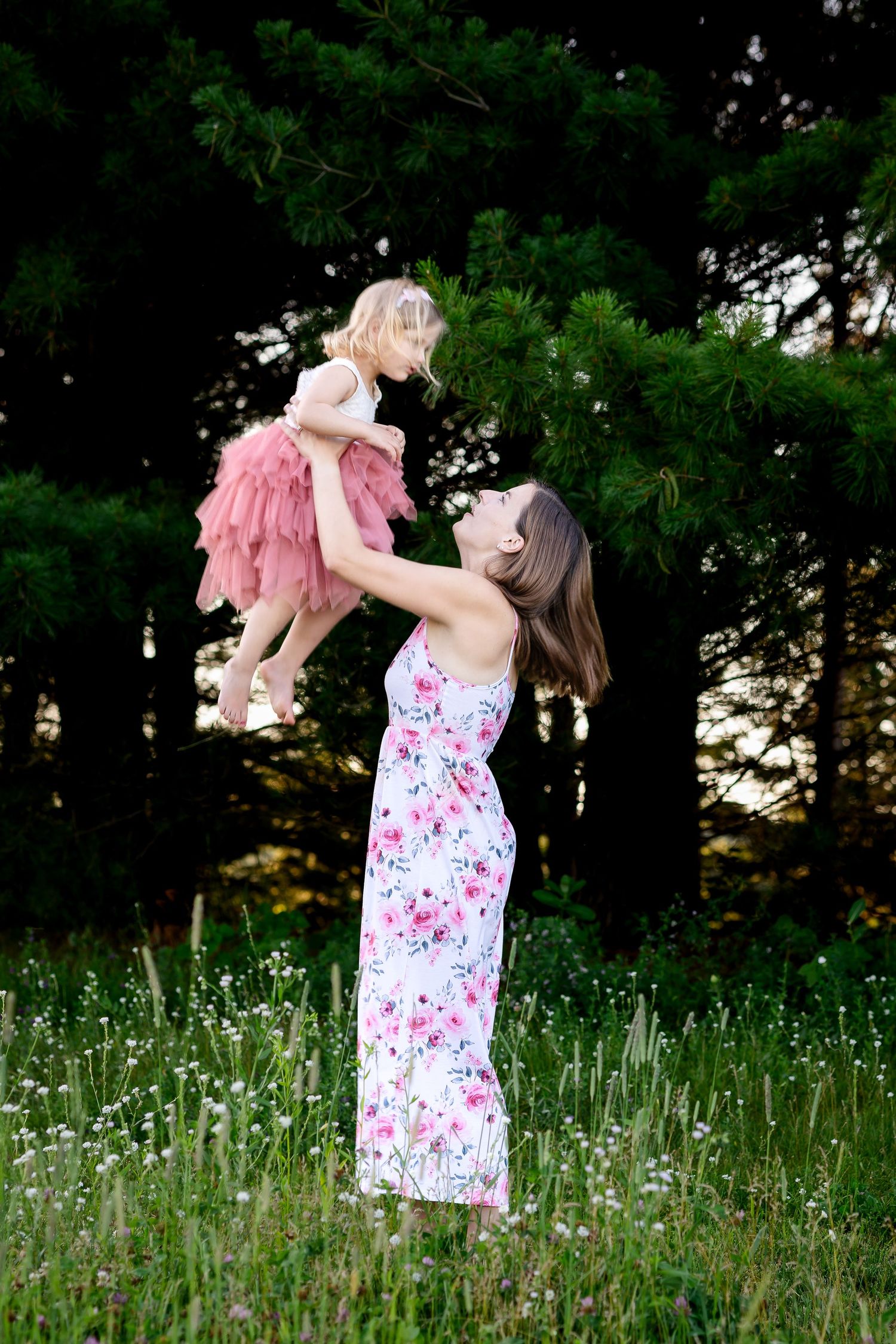 Parent lifts child in pink dress while standing in wildflower meadow at sunset.