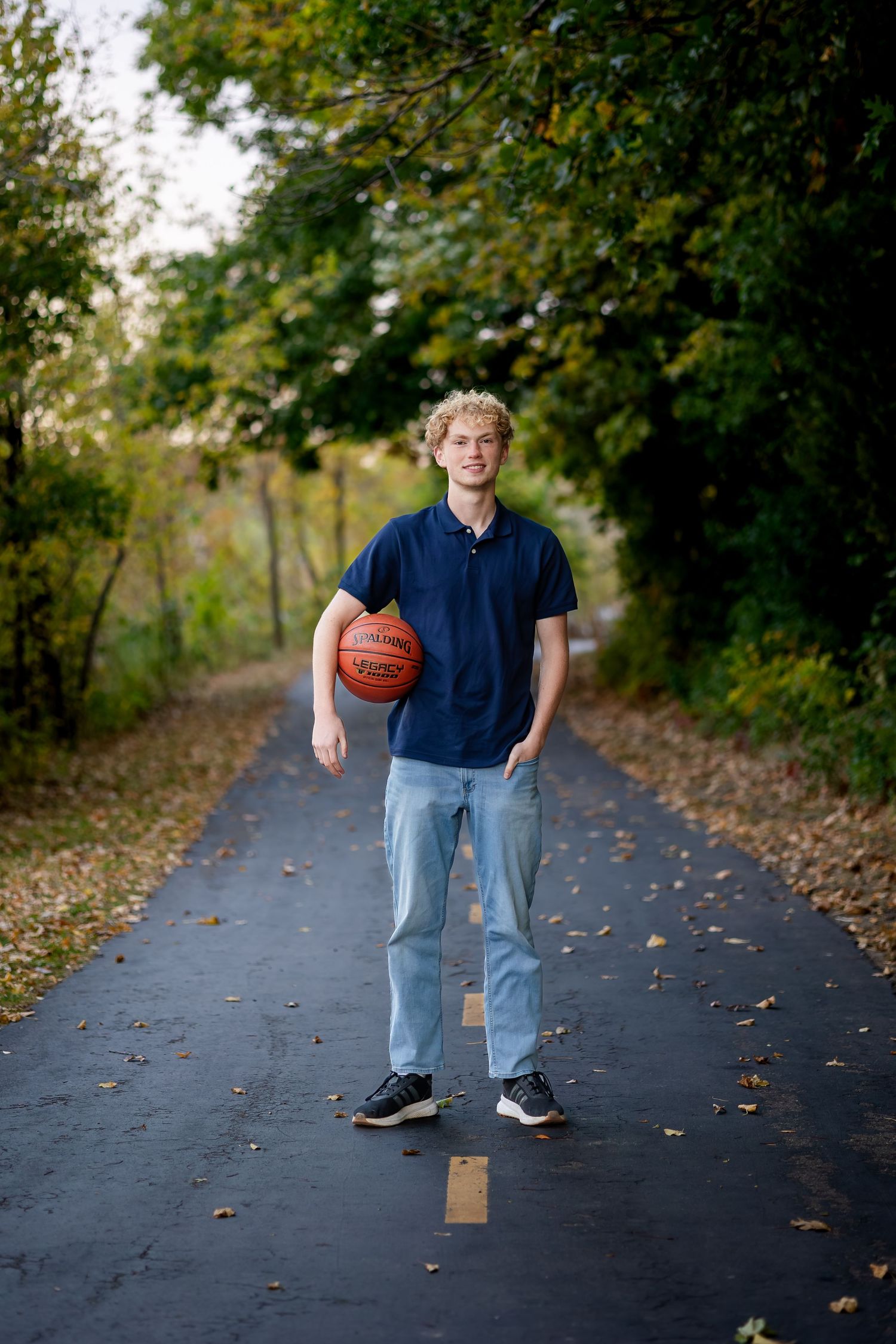 A person stands on a tree-lined path holding a basketball while wearing a navy polo shirt and light jeans.