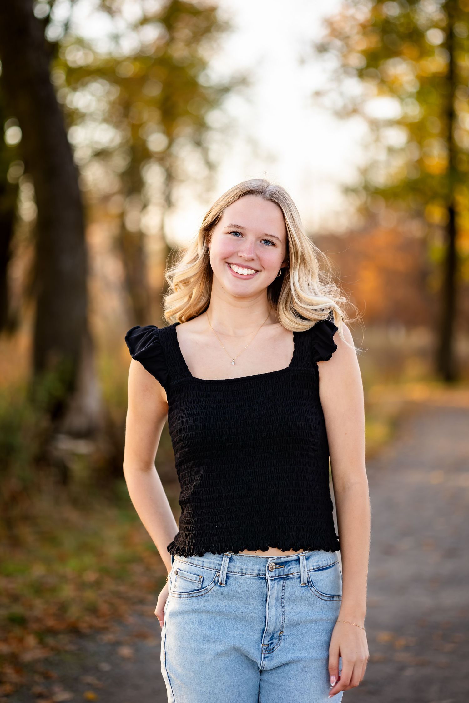 A person in a black top and light denim jeans poses outdoors on an autumn path with fallen leaves.