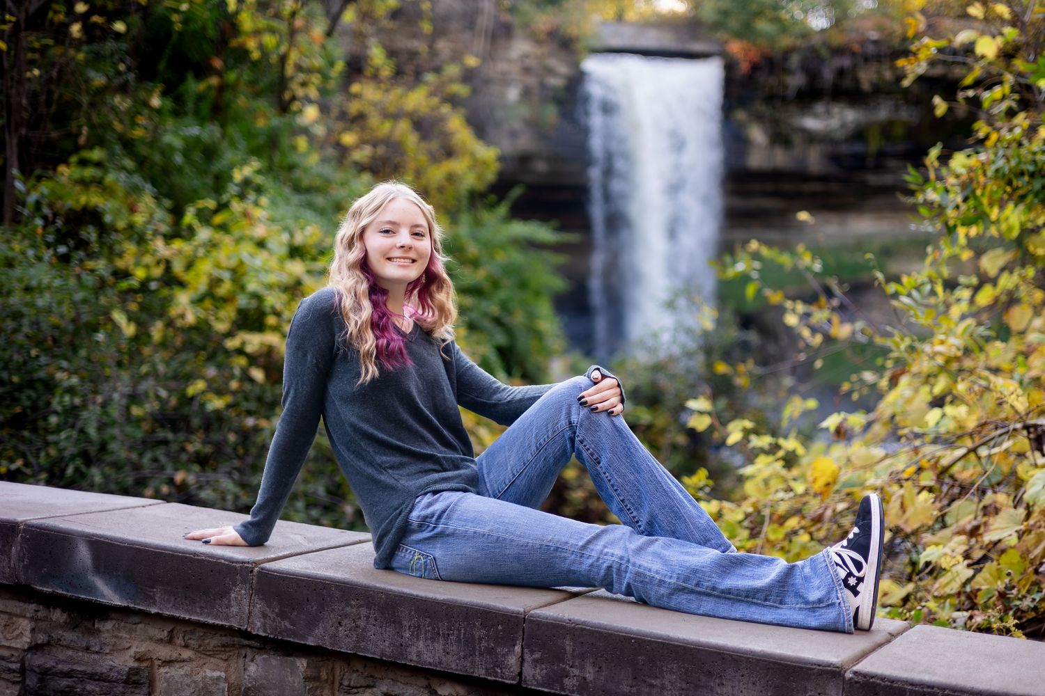 Person in jeans and dark top lounging on wooden railing with autumn foliage and waterfall in background.