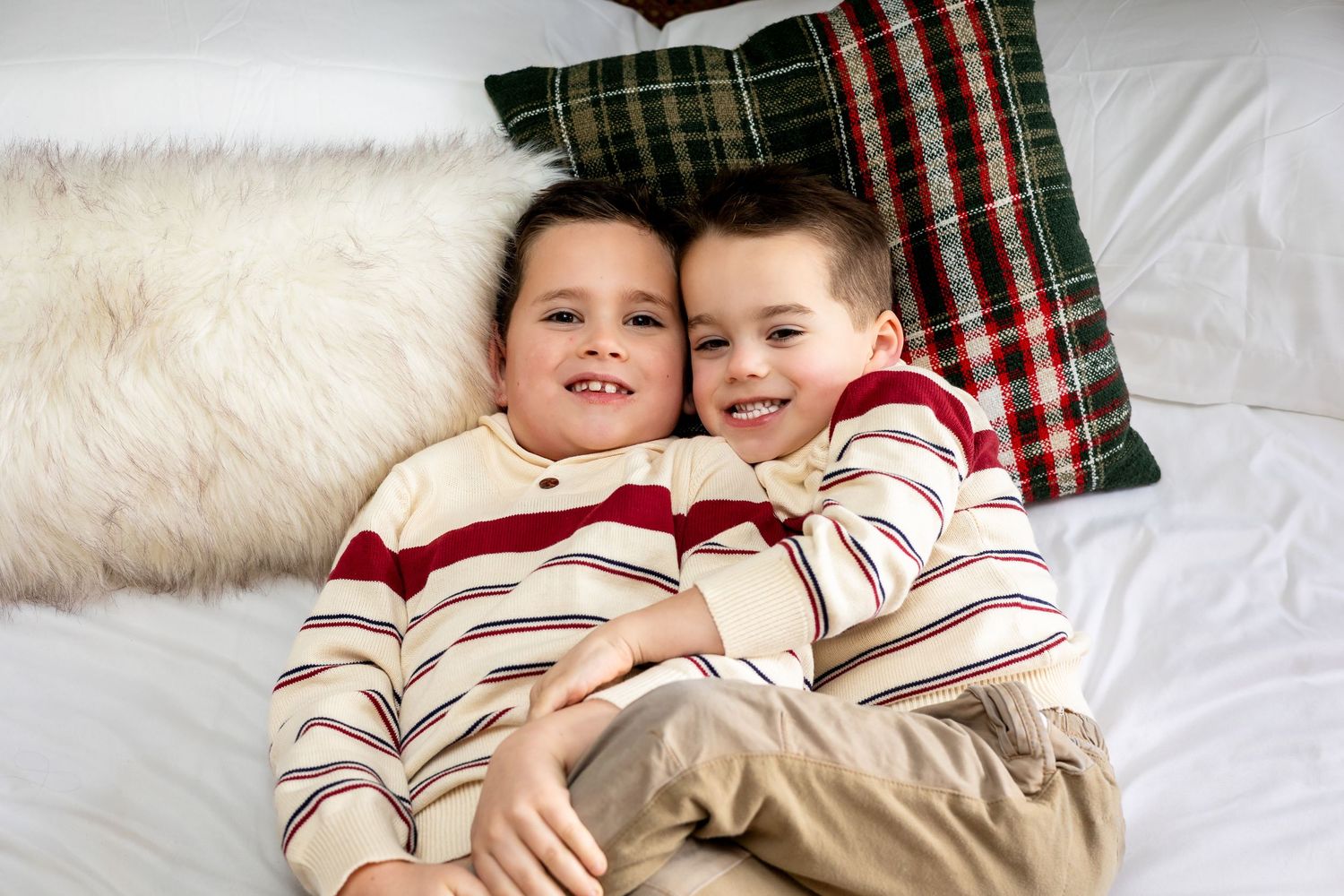 Two young boys in matching striped holiday pajamas cuddle together on a white bed with plaid pillows.