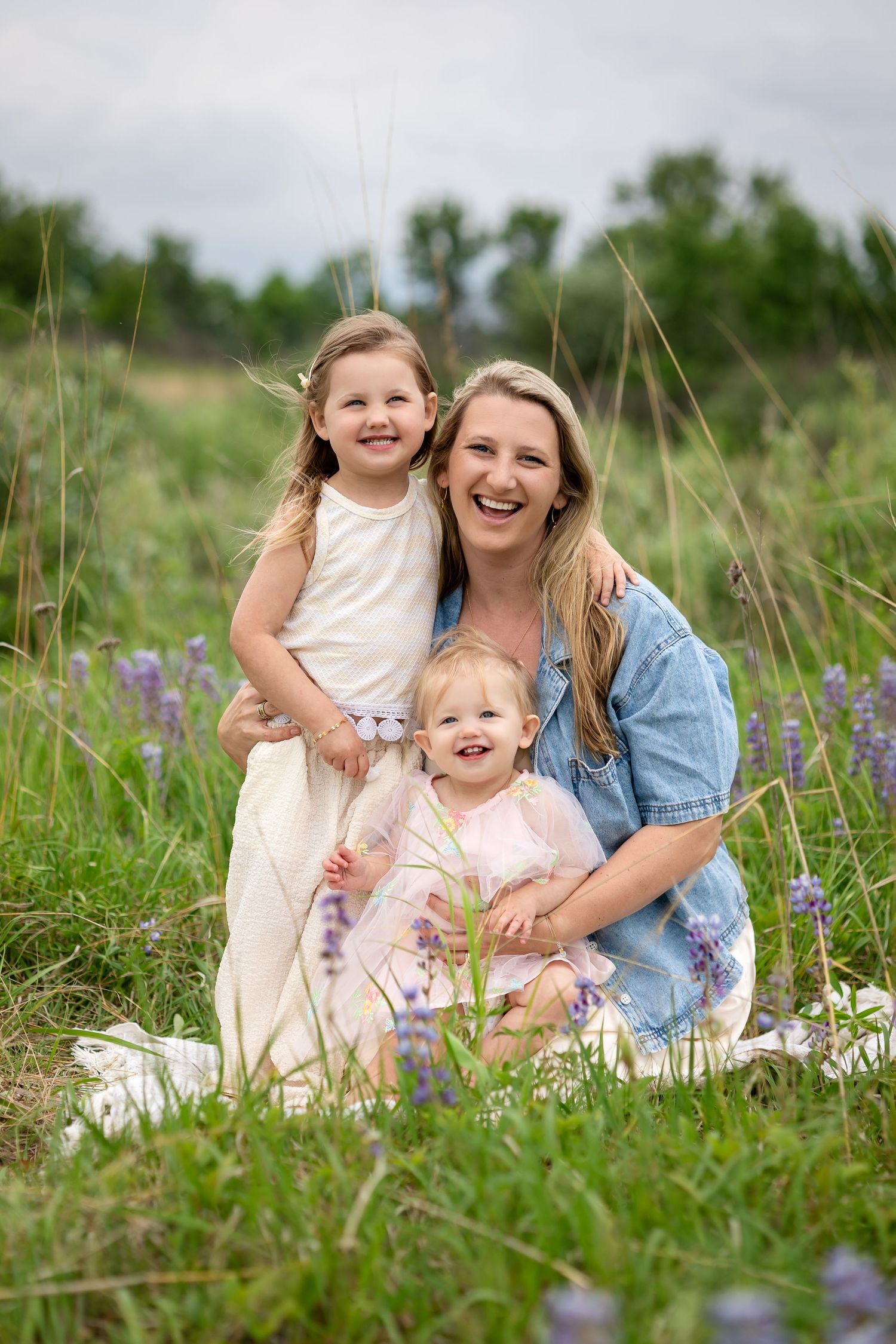 A smiling mother poses outdoors in a green field with her two young daughters wearing white and pink dresses.