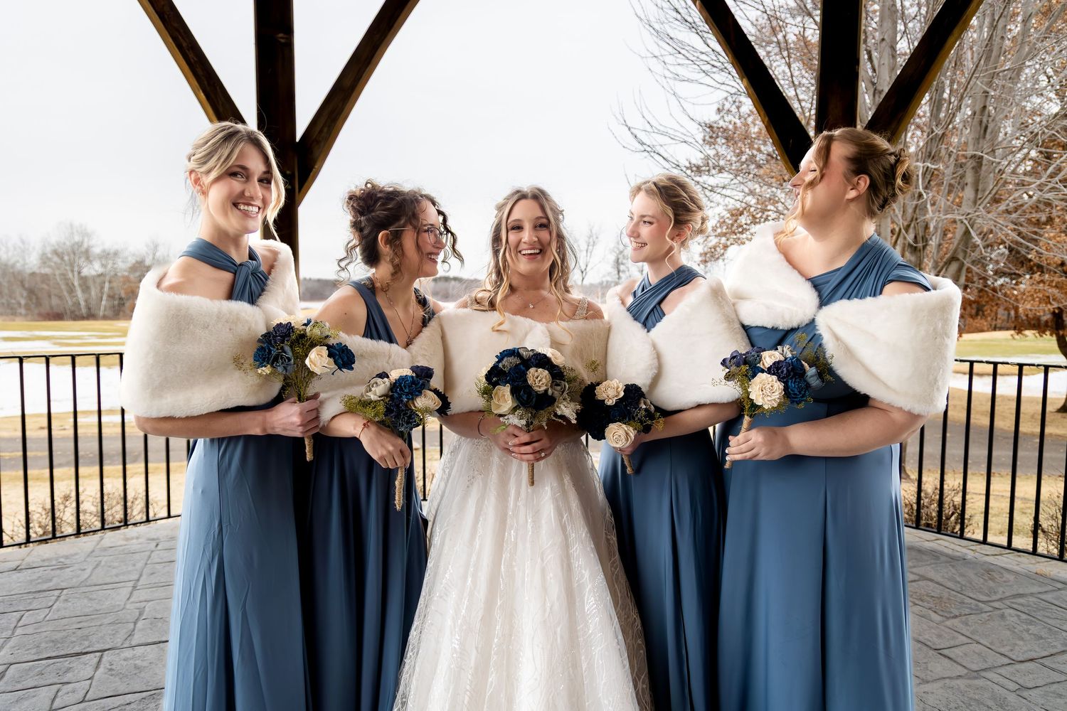 Bride poses with four bridesmaids in dusty blue dresses and white fur wraps holding navy and white bouquets.